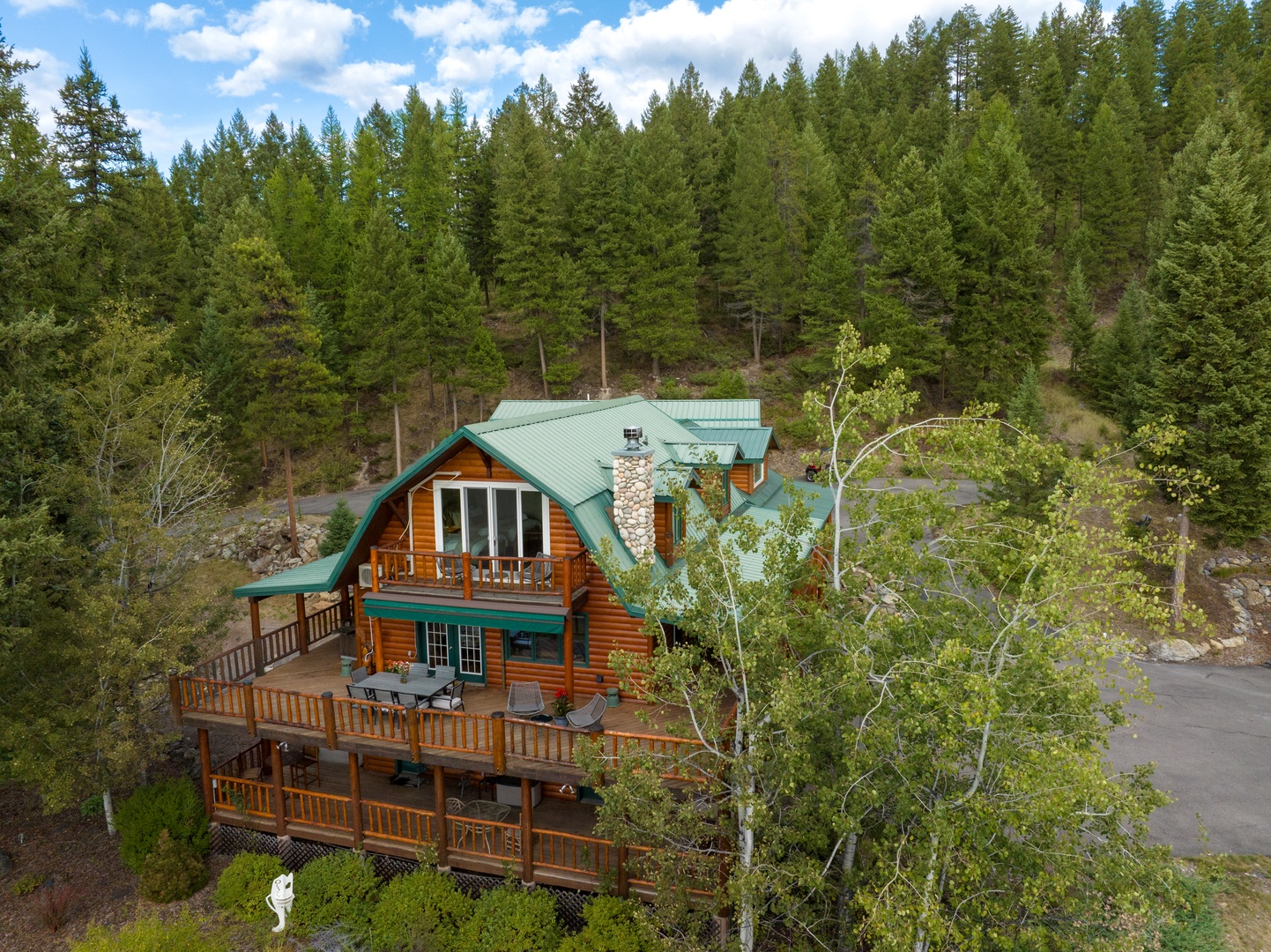 Charming log cabin nestled in a pristine forest setting with towering evergreens and multiple decks. The distinctive green metal roof and natural wood siding blend beautifully with the surrounding wilderness landscape.