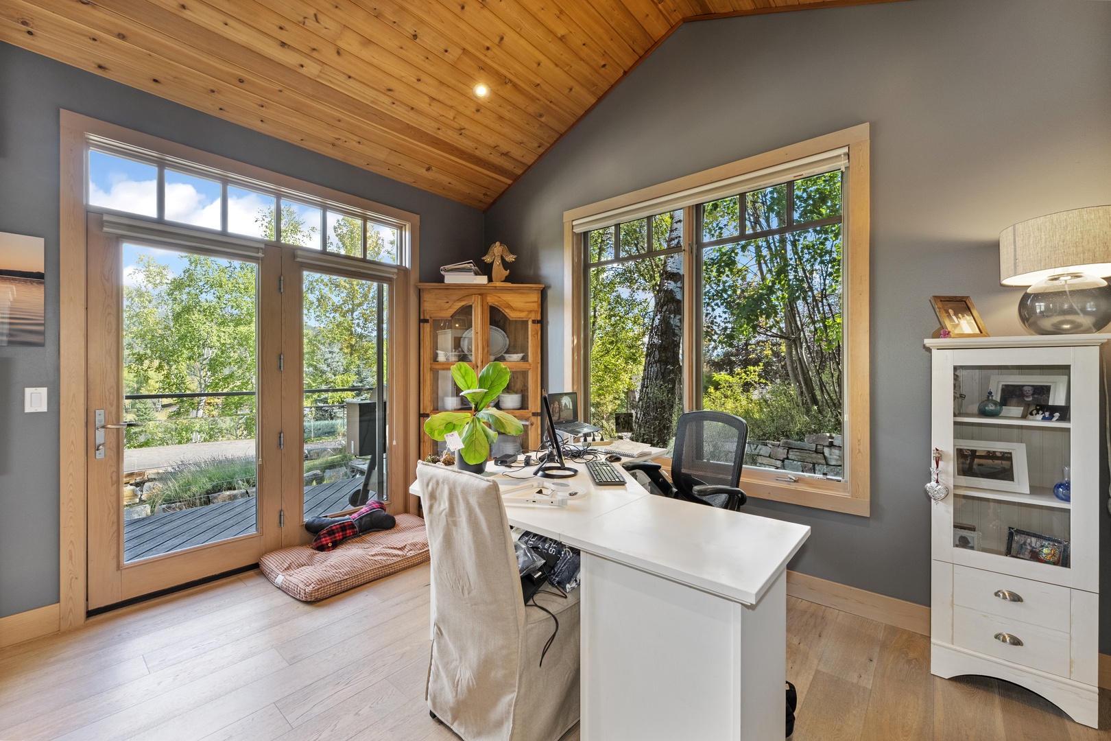 Your bright home office overlooks lush trees through expansive windows, letting natural light pour in while you work under the warm glow of vaulted wood ceilings.