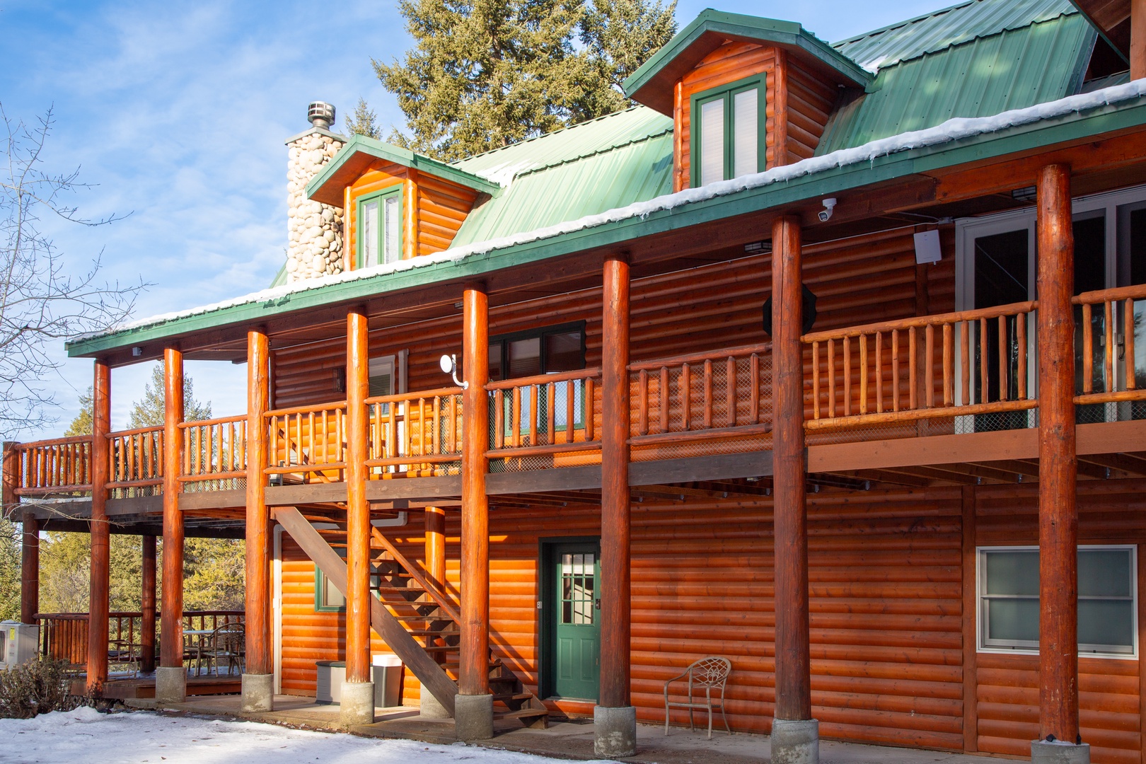 A handsome log cabin with green metal roofing and wraparound deck, set among mature trees with snow lightly dusting the ground and steps.