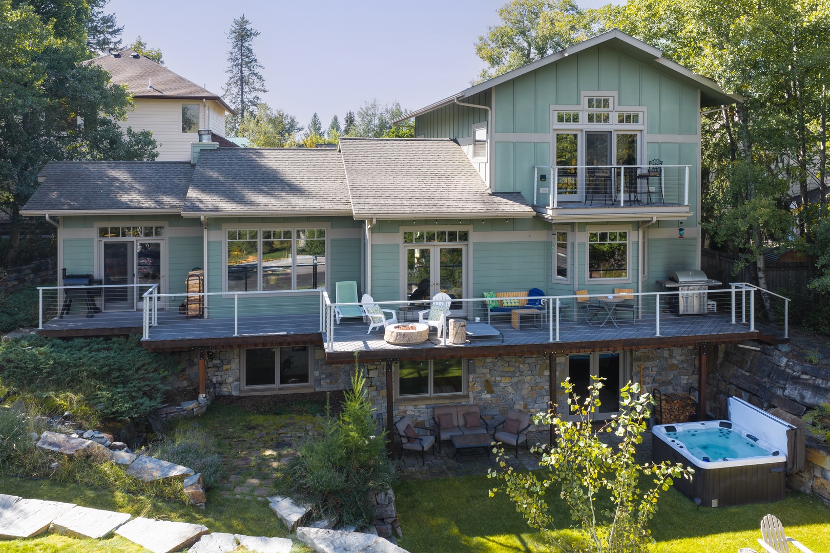 A charming multi-level retreat set among towering trees, featuring a spacious deck for gathering, a stone-walled lower patio, and an inviting hot tub nestled in the backyard.
