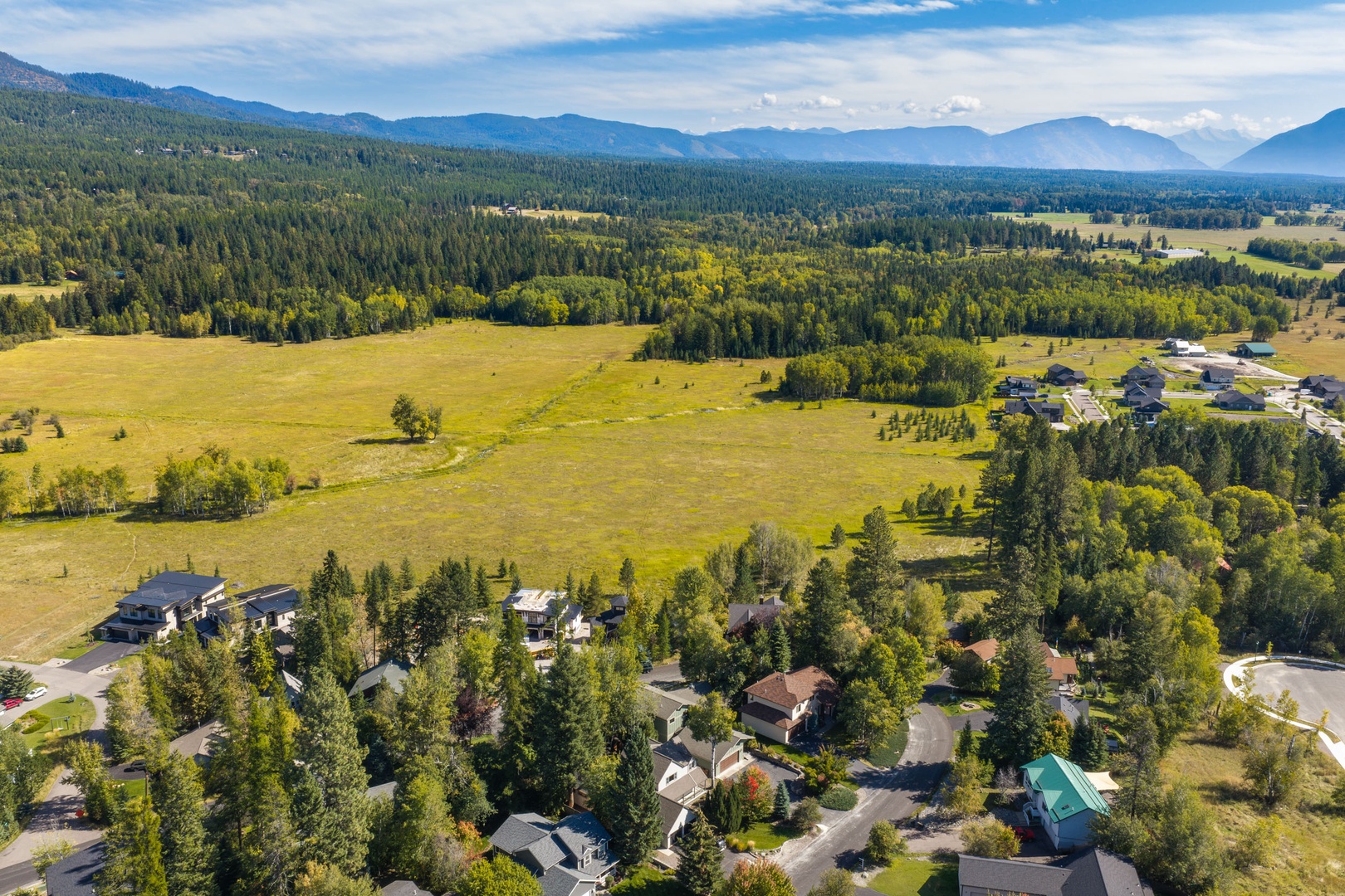 An aerial view captures a peaceful mountain valley neighborhood surrounded by sprawling forests, open meadows, and dramatic peaks in the distance—part of the scenic landscape near the property.