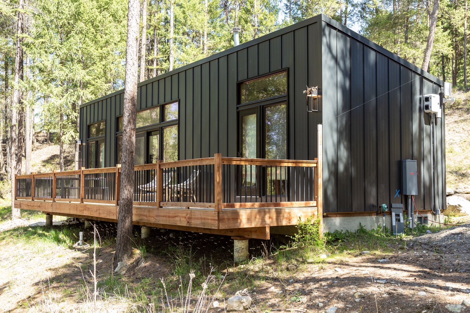 A modern retreat nestled among tall pines, featuring dark metal siding and a spacious wraparound deck perfect for enjoying the peaceful forest setting.