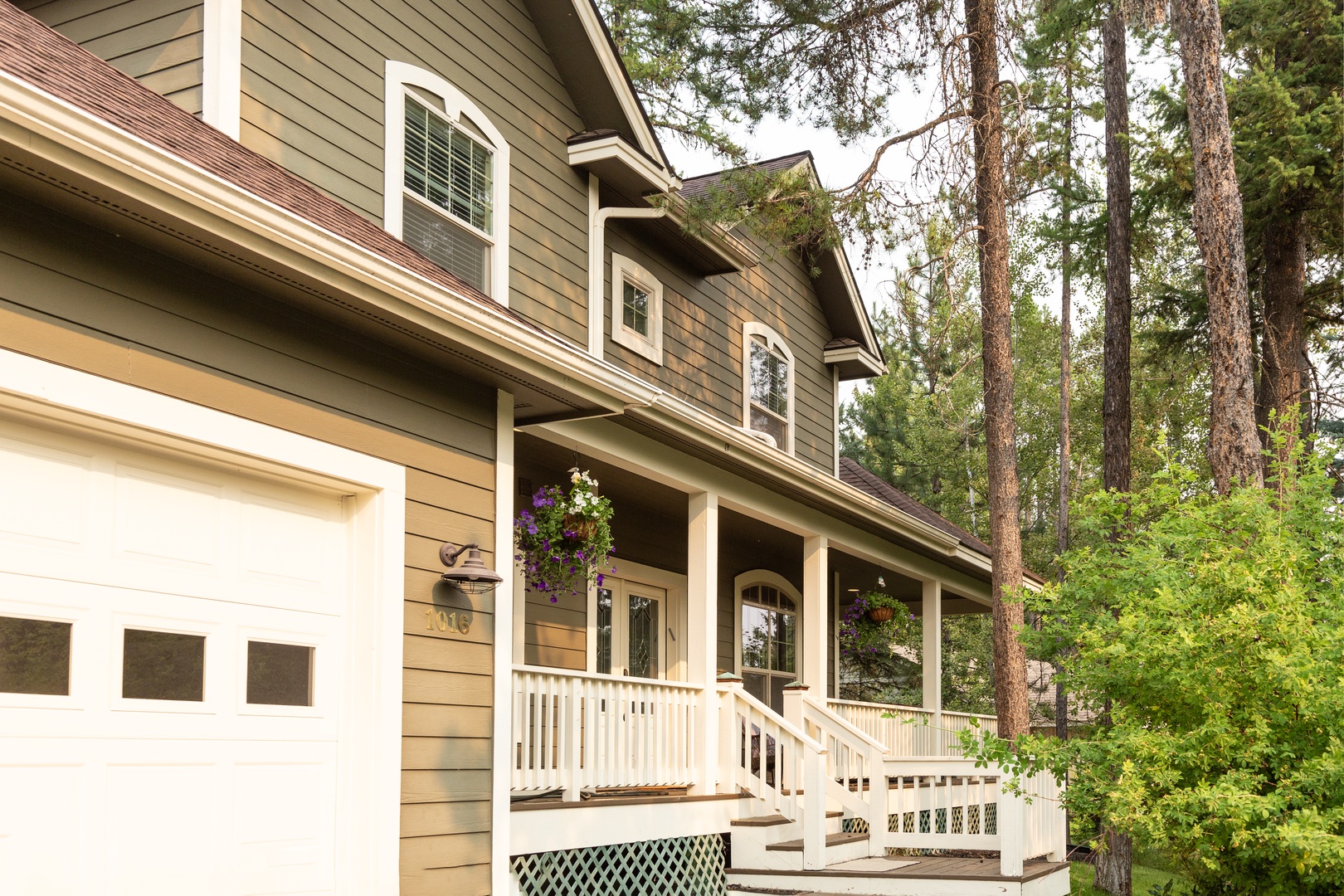 A welcoming two-story property nestled in a peaceful wooded setting, framed by towering pines and lush greenery. The covered front porch with white railings and hanging flower baskets invites a relaxing arrival.