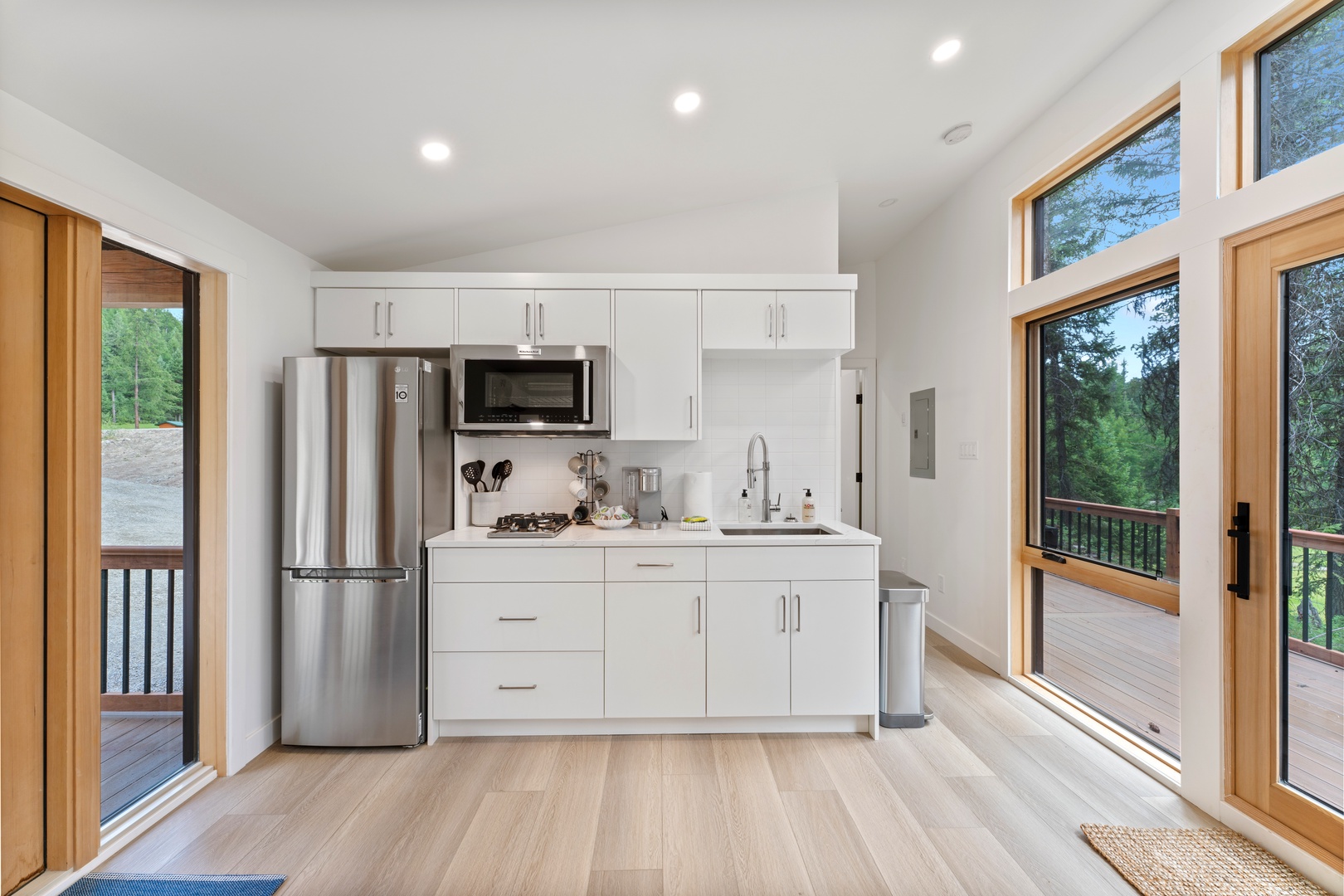 Your modern kitchenette features sleek white cabinets, stainless steel appliances, and stunning forest views through oversized windows that flood the space with natural light.