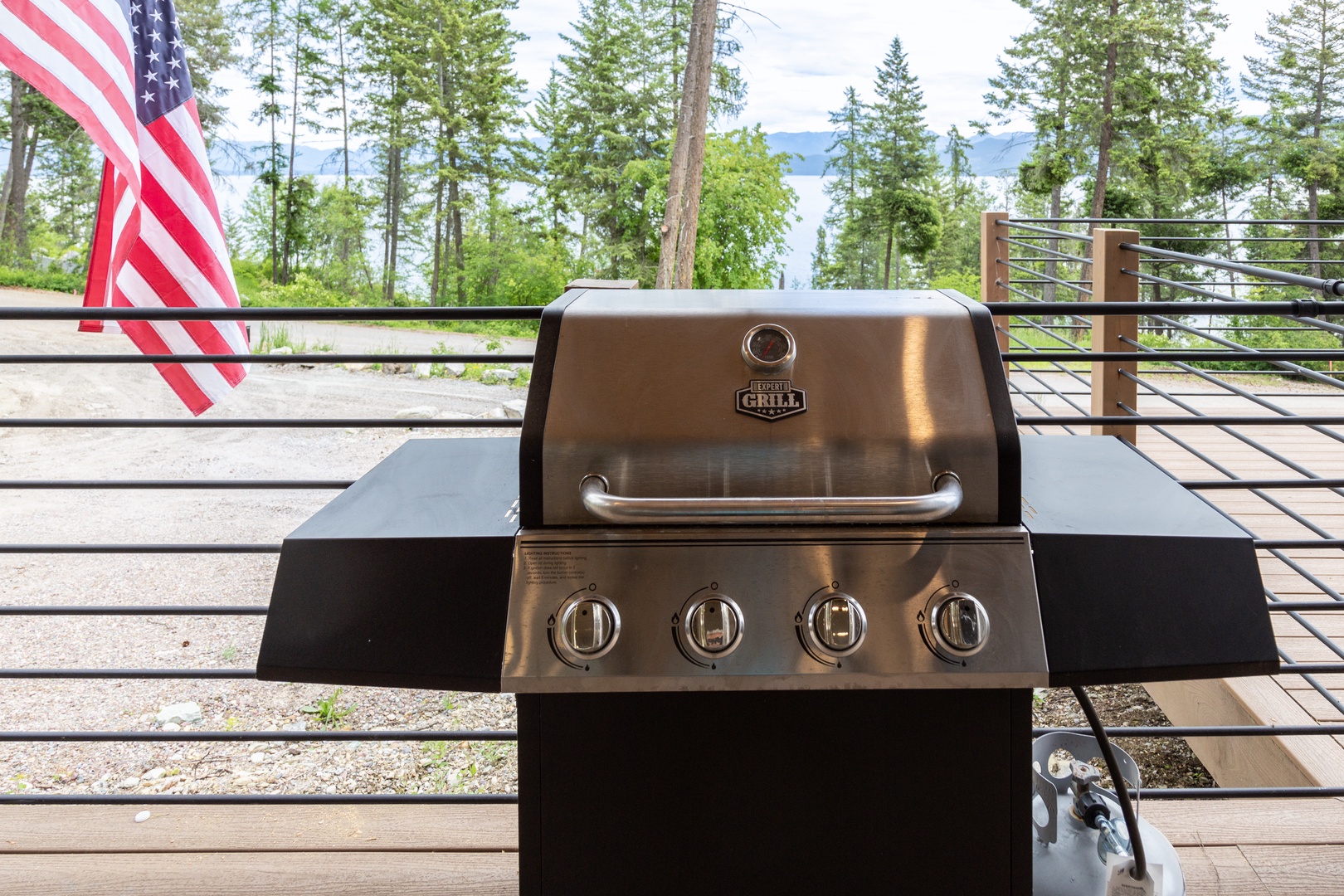 A stainless steel grill is situated on an outdoor deck, overlooking forested landscape and lake views, with an American flag visible nearby.