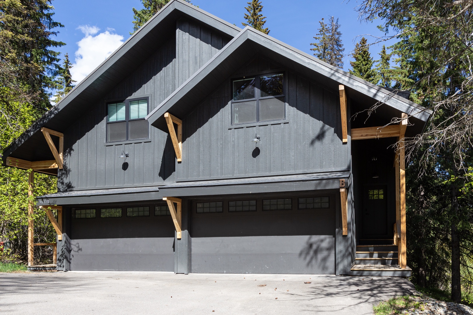 Modern mountain retreat with striking charcoal facade and warm wood accents, nestled among towering evergreens with convenient parking and welcoming entrance.