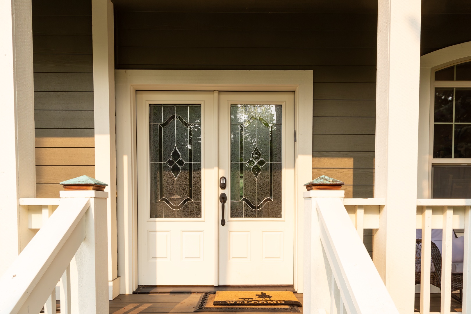 Warm light welcomes you at the double-door entrance, featuring elegant glass panels and a covered porch with classic white railings—your retreat awaits just beyond this charming threshold.