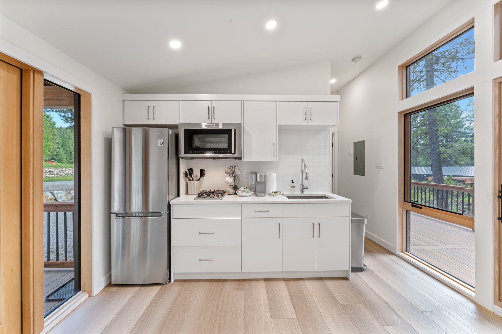 Your modern kitchenette features sleek white cabinets, stainless steel appliances, and beautiful forest views through large windows.