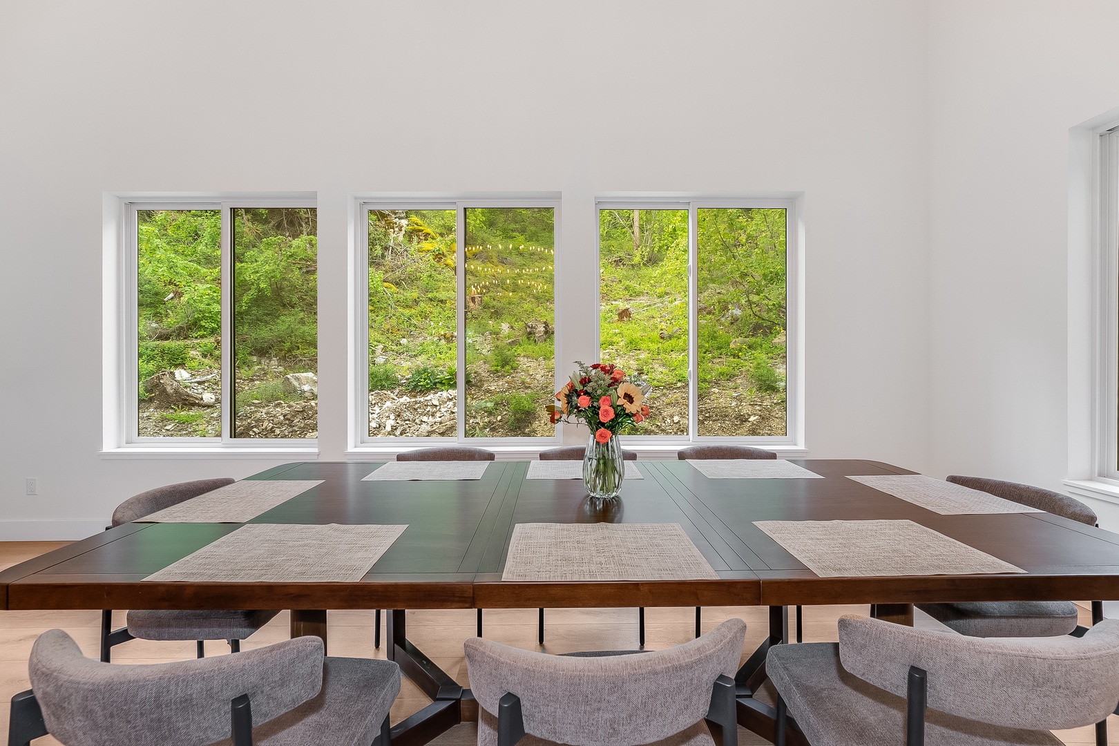 Gather with loved ones at this inviting dining table, set with fresh flowers and surrounded by windows that frame lush greenery—your backdrop for memorable meals together.