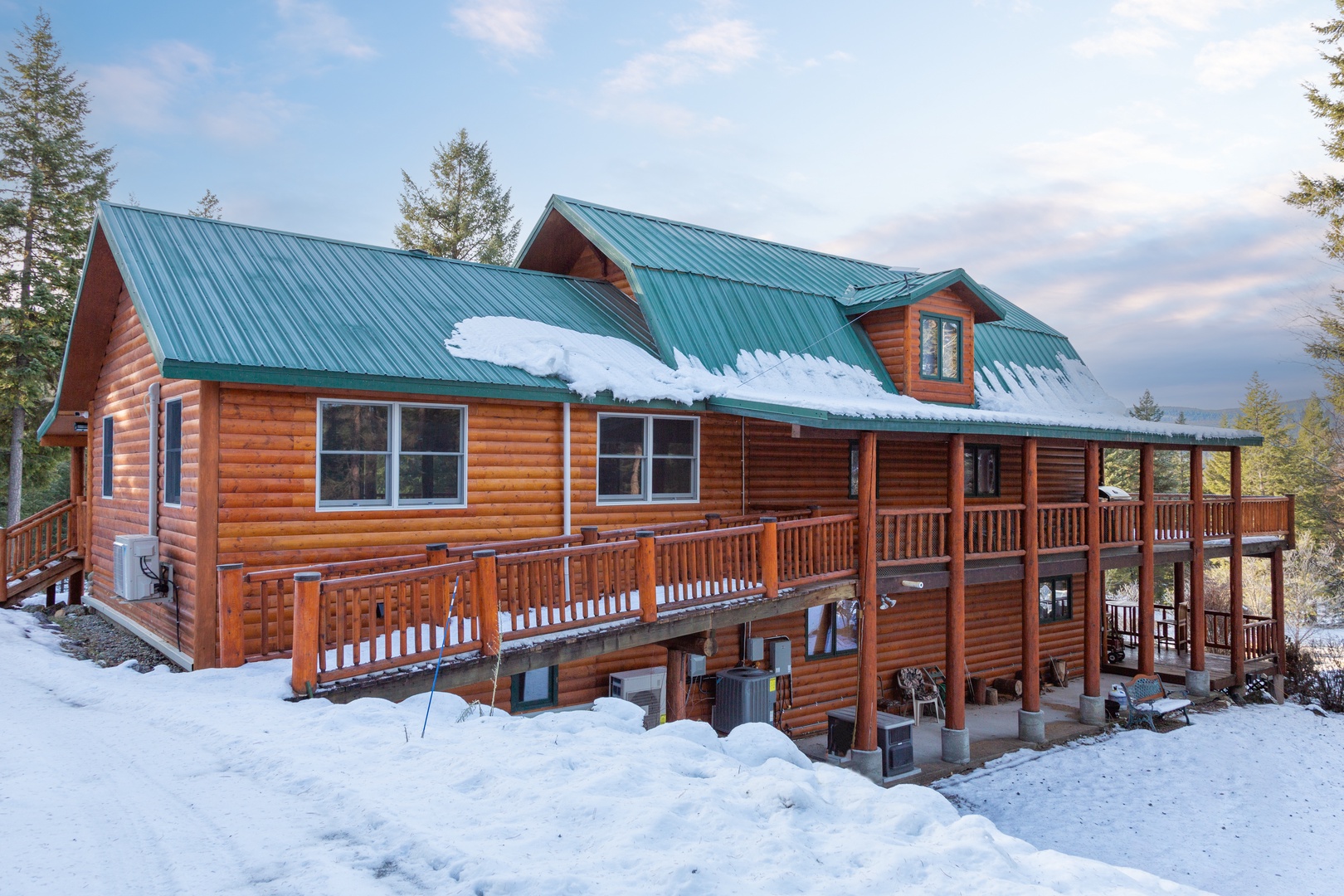 A spacious mountain lodge with warm log siding, green metal roof, and expansive decks overlooking a forested winter landscape—a serene retreat amid pine trees and snow.