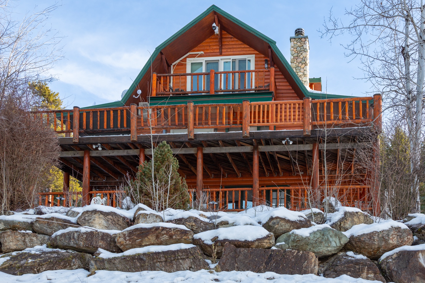 A charming log cabin with a distinctive green roof and stone chimney rises above a natural rock foundation, surrounded by snow-dusted trees and winter scenery in this peaceful mountain setting.
