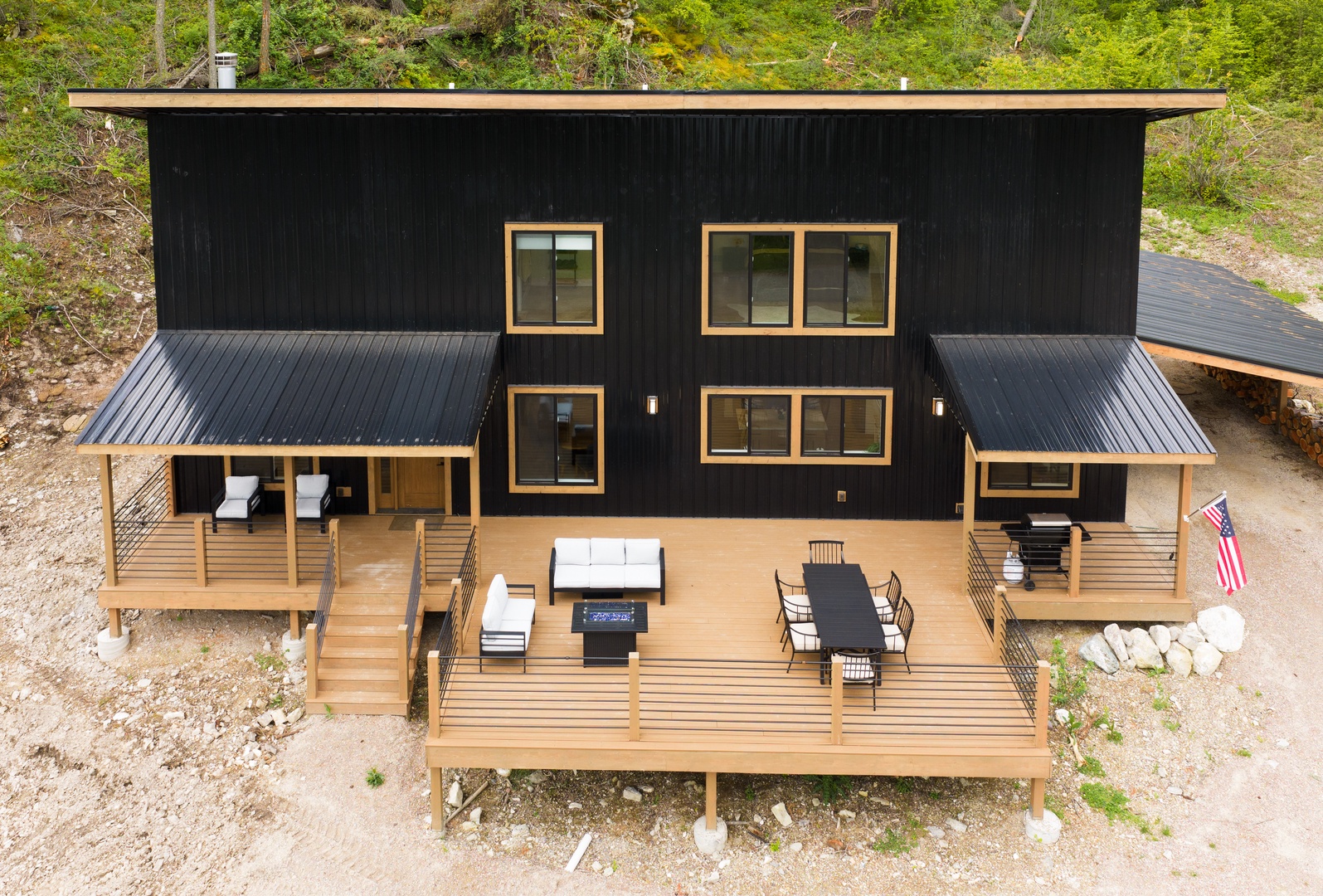 A striking modern cabin with black facade and warm wood accents, set on a wooded hillside, featuring an expansive multi-level deck furnished for dining and relaxation.