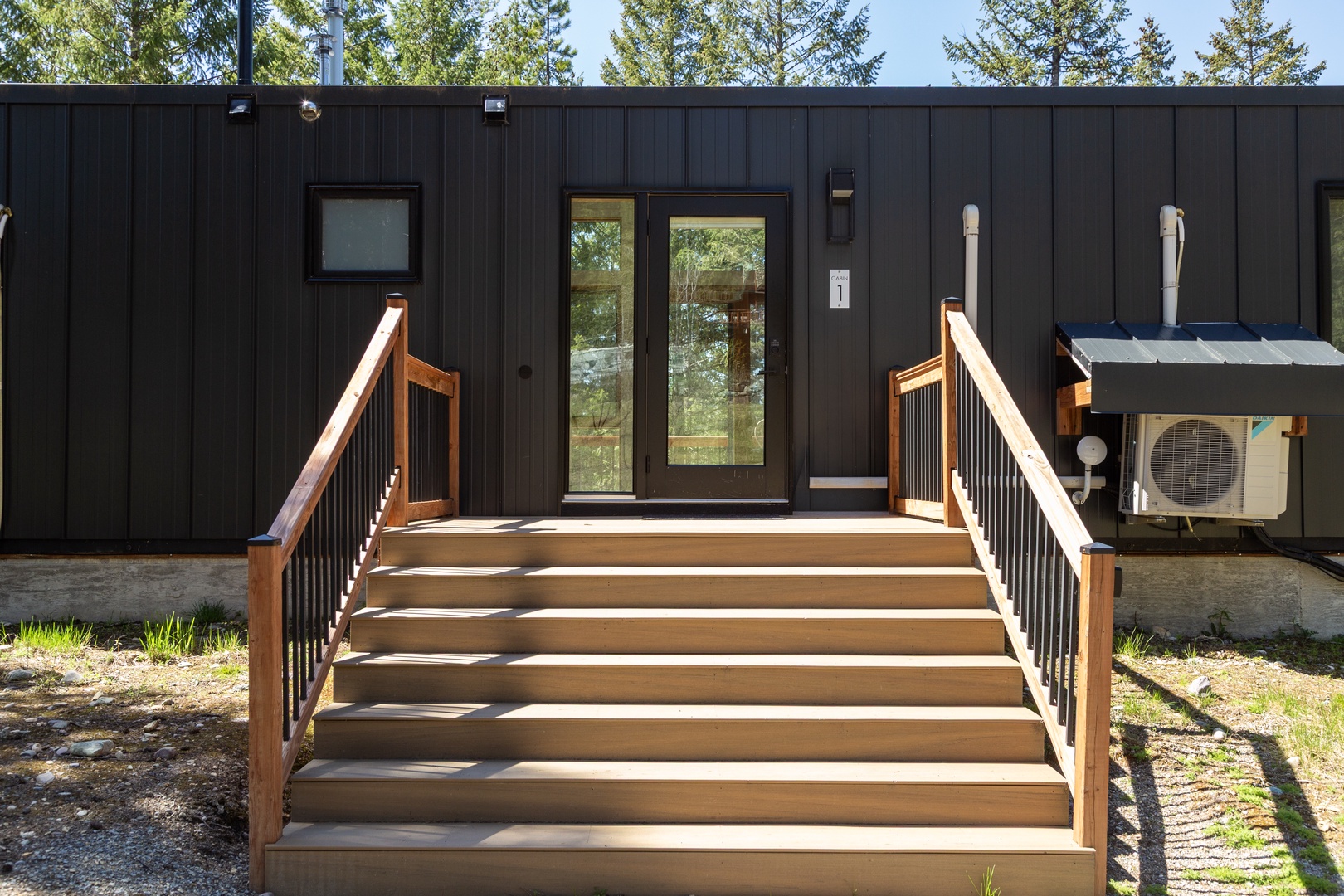 The modern black exterior and warm wood staircase mark the welcoming entrance to the property, surrounded by tall pines in a peaceful natural setting.