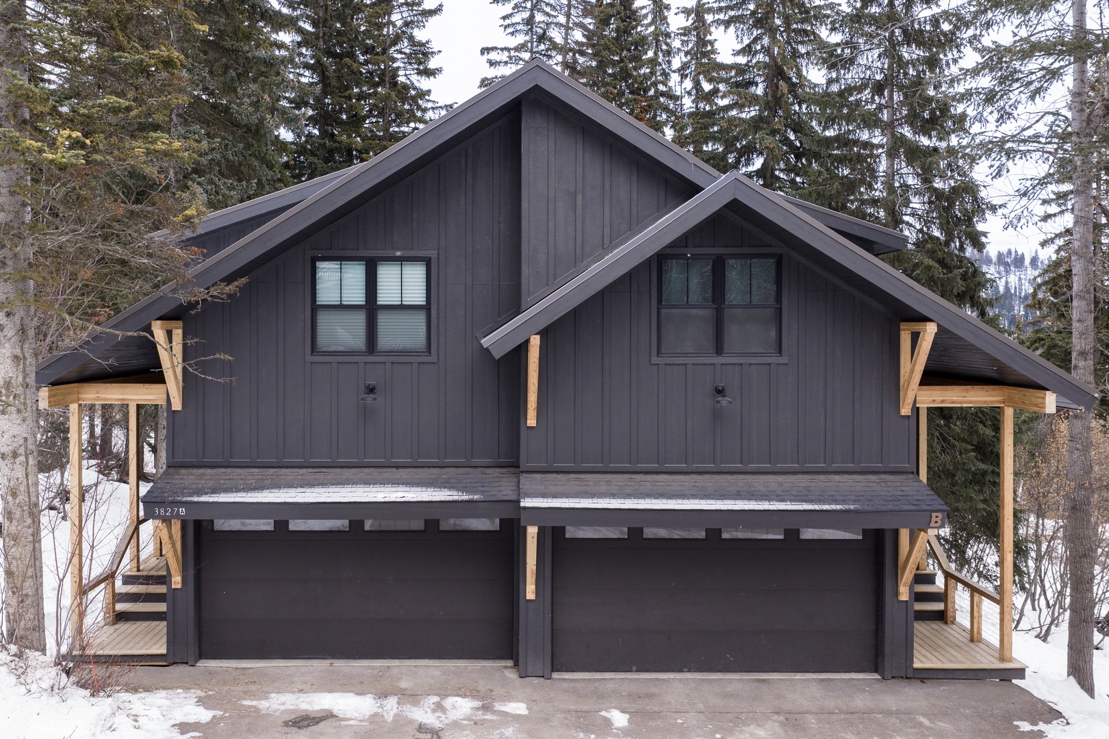 Modern mountain duplex nestled among towering evergreens, featuring dark wood siding and contemporary alpine architecture in a peaceful winter setting.