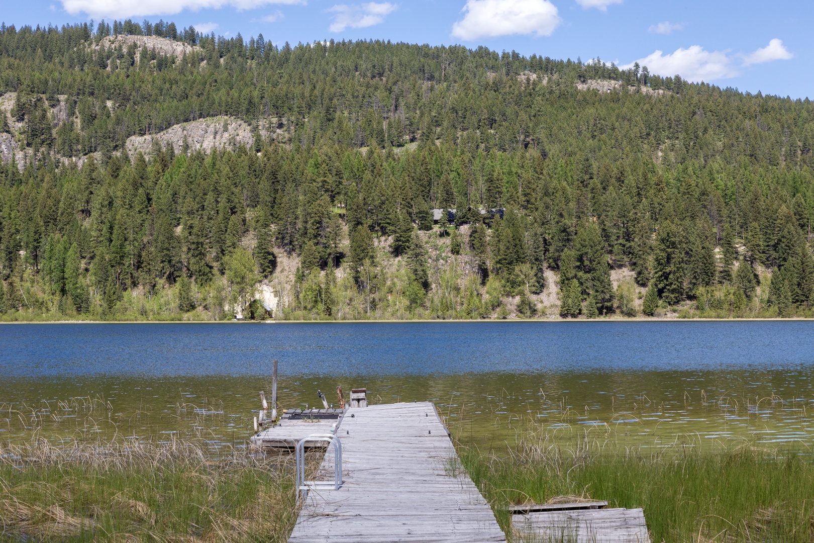 A peaceful lakefront setting features a private dock stretching into calm waters, framed by forested mountainsides and clear skies—an idyllic natural escape in the surrounding area.