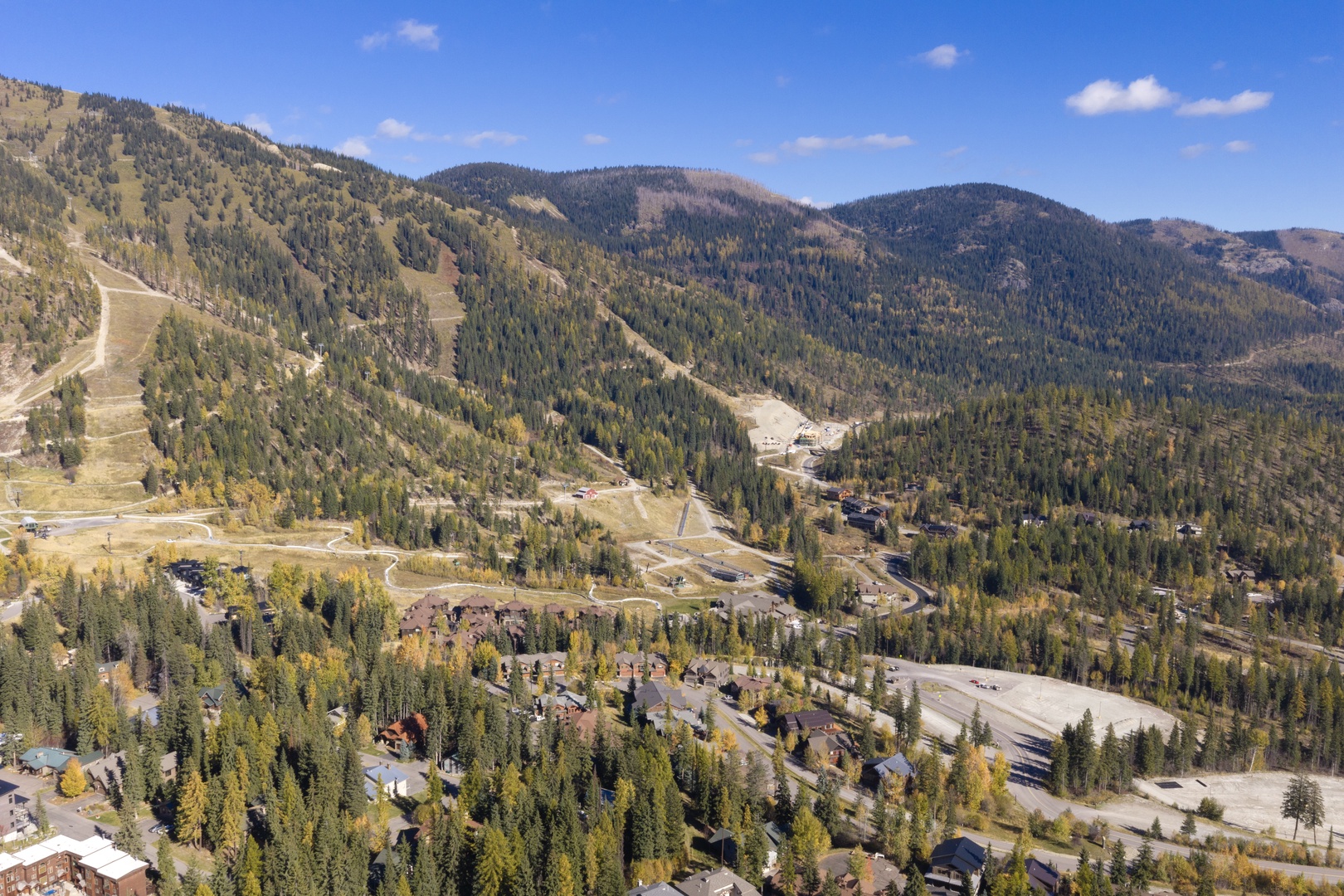 Aerial view of a mountain valley community nestled among evergreen forests and seasonal foliage, with ski slopes and winding roads visible throughout the landscape.