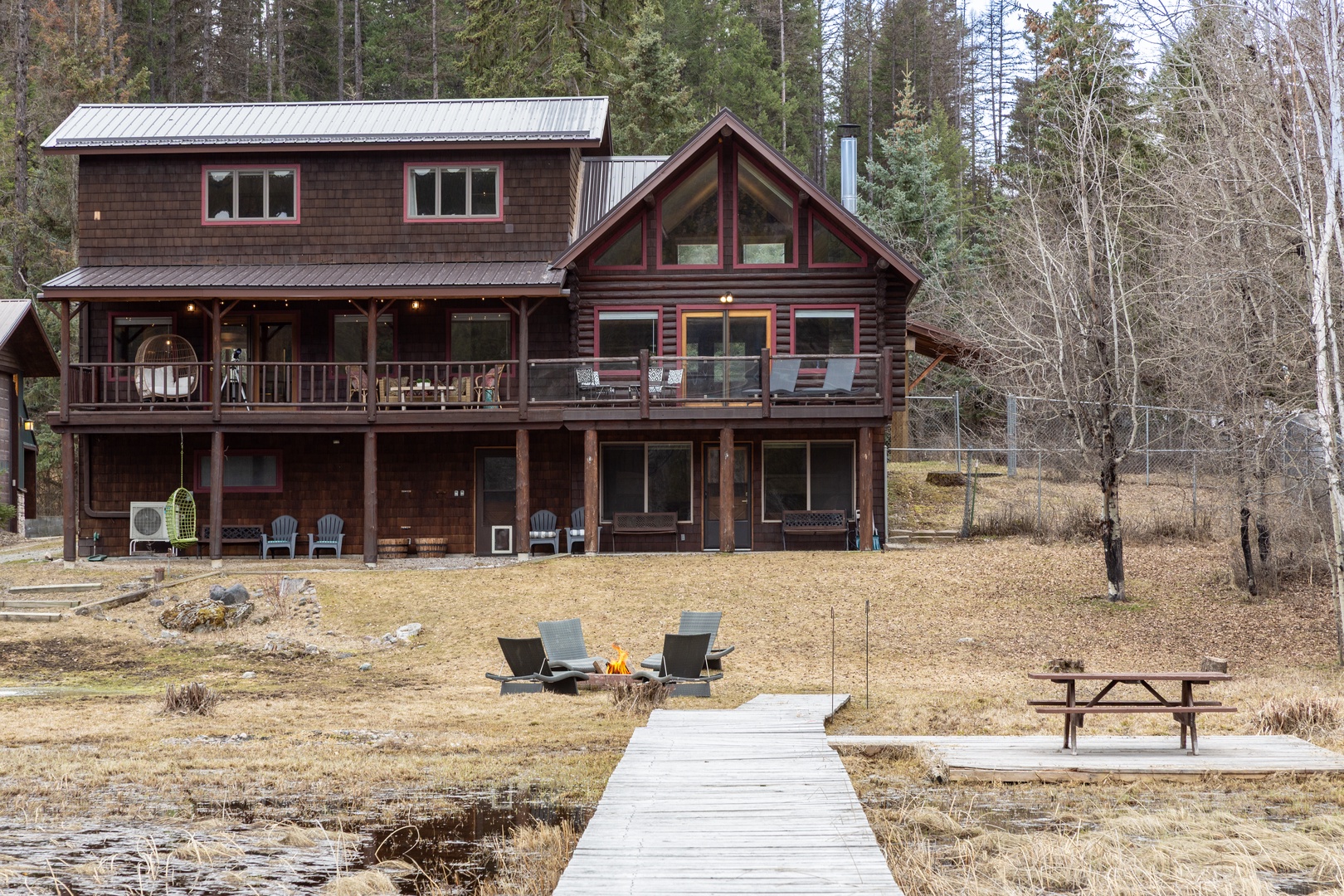 Rustic lakeside cabin with wooden dock, fire pit area, and picnic table surrounded by peaceful forest setting.