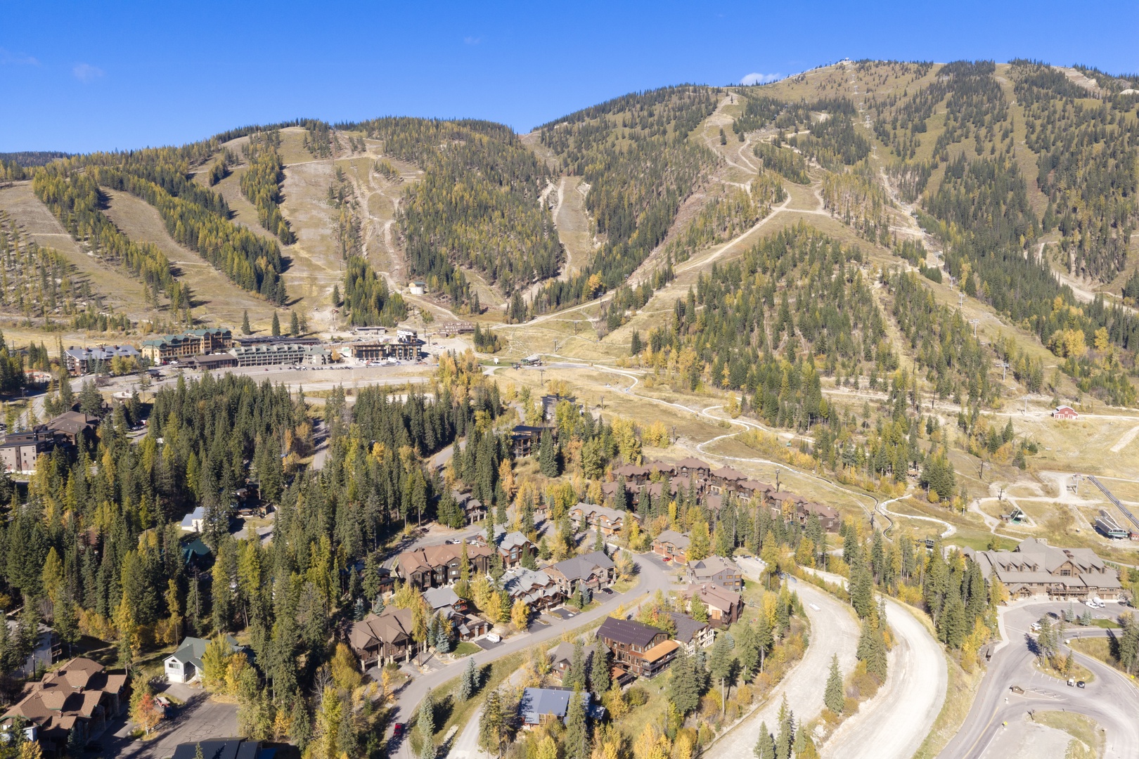An aerial view showcases a mountain resort community nestled among evergreen forests and rolling ski slopes, with winding roads connecting rustic lodges to the base area.