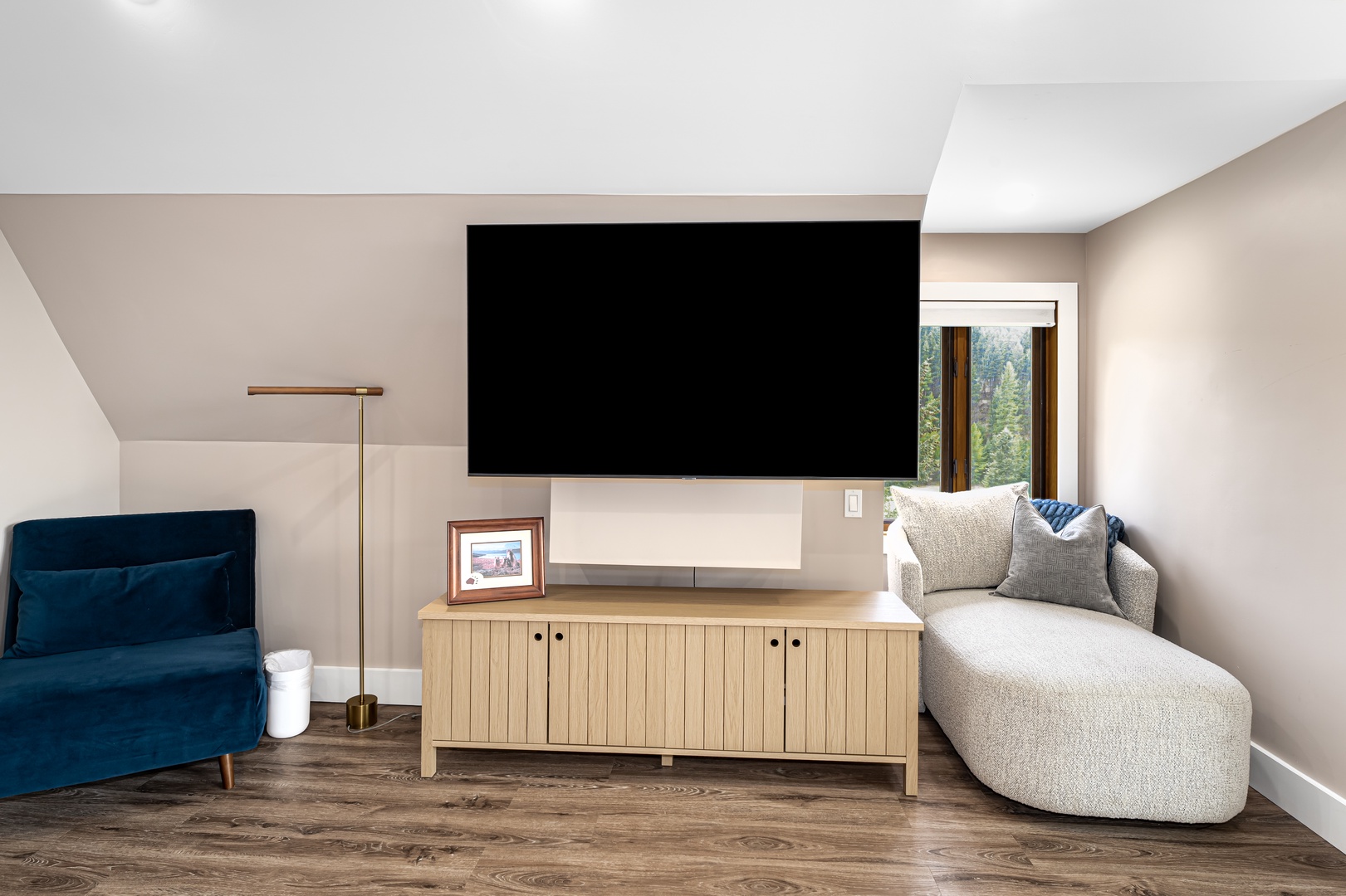 Unwind in your stylish living area with a large flat-screen TV and cozy seating, framed by natural light and forest views through the window.