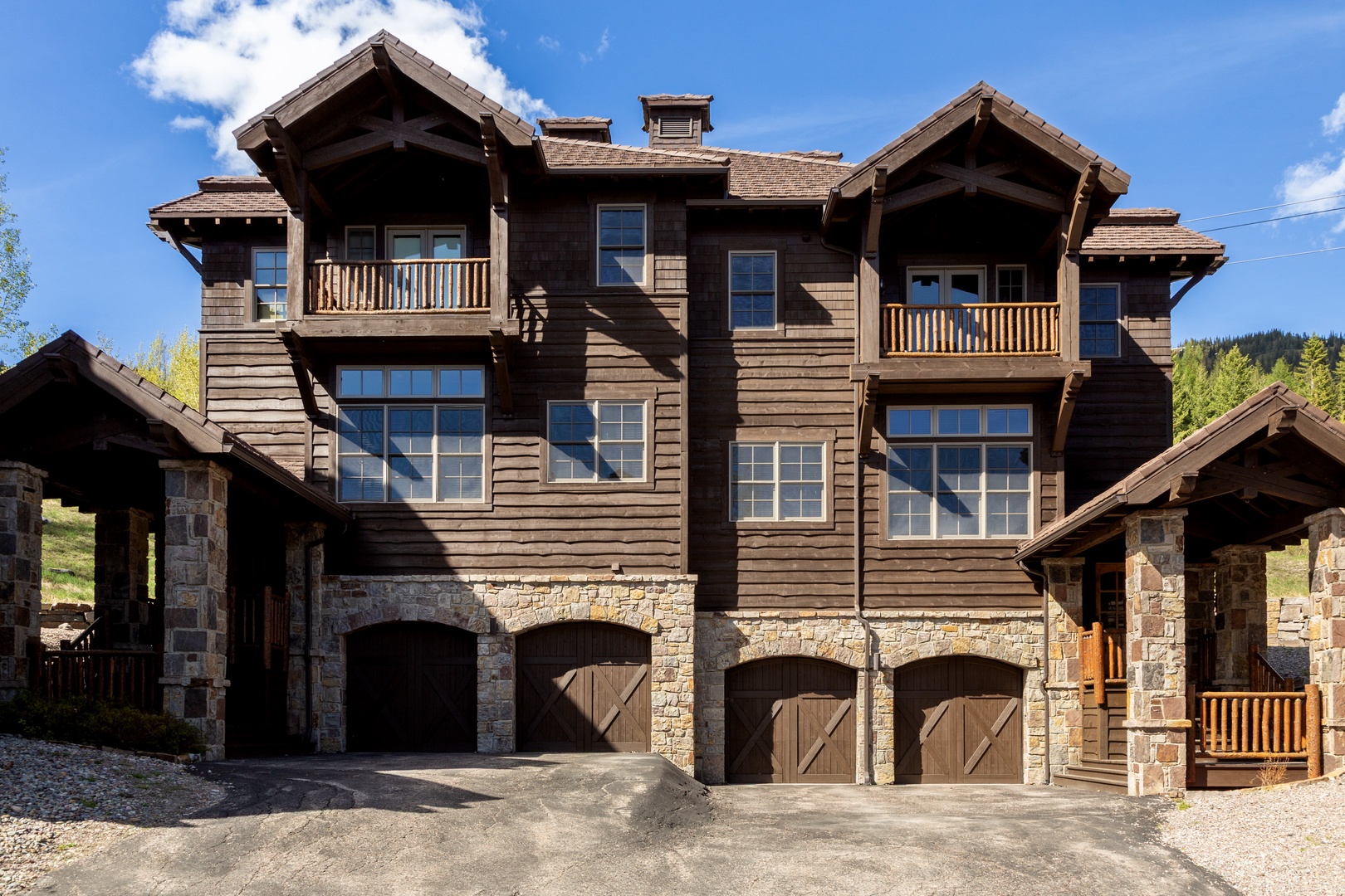 A striking alpine-inspired mountain residence featuring rustic stone and wood siding, multiple private balconies, and arched garage doors set against forested hillsides under clear blue skies.