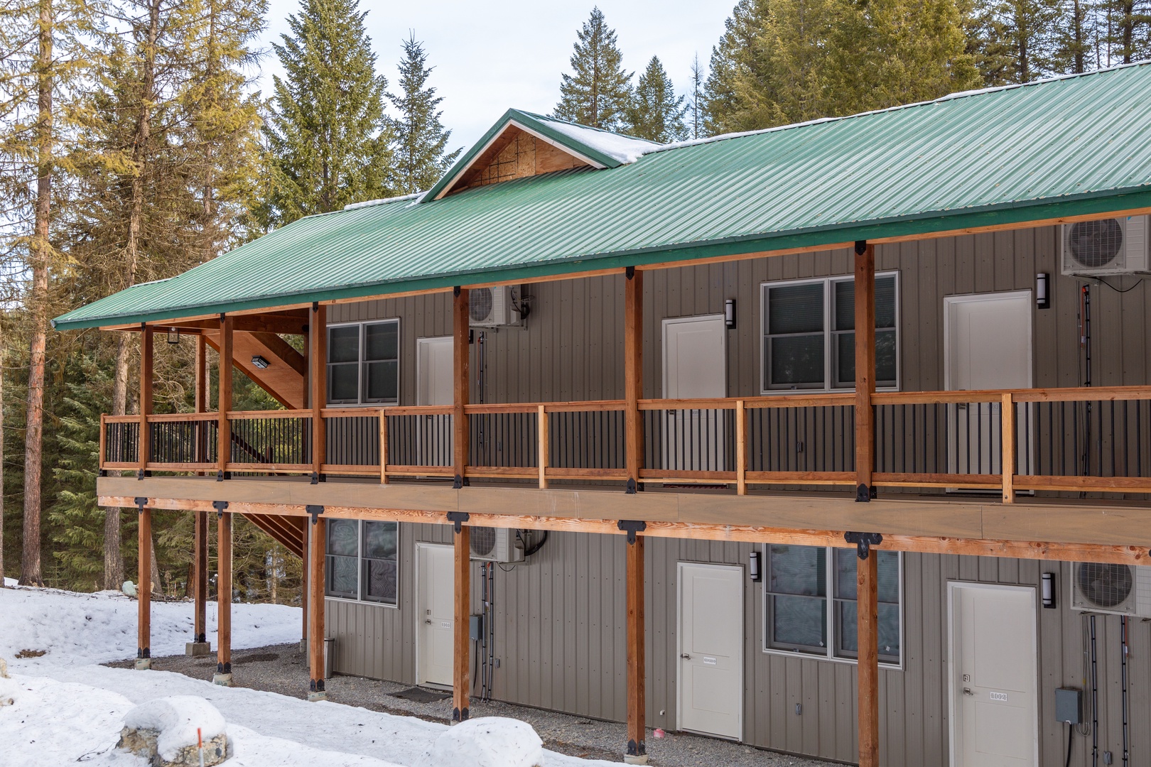 A two-story mountain retreat with green metal roofing nestled among towering evergreens, featuring individual unit entrances and covered balcony access in a peaceful forested setting.