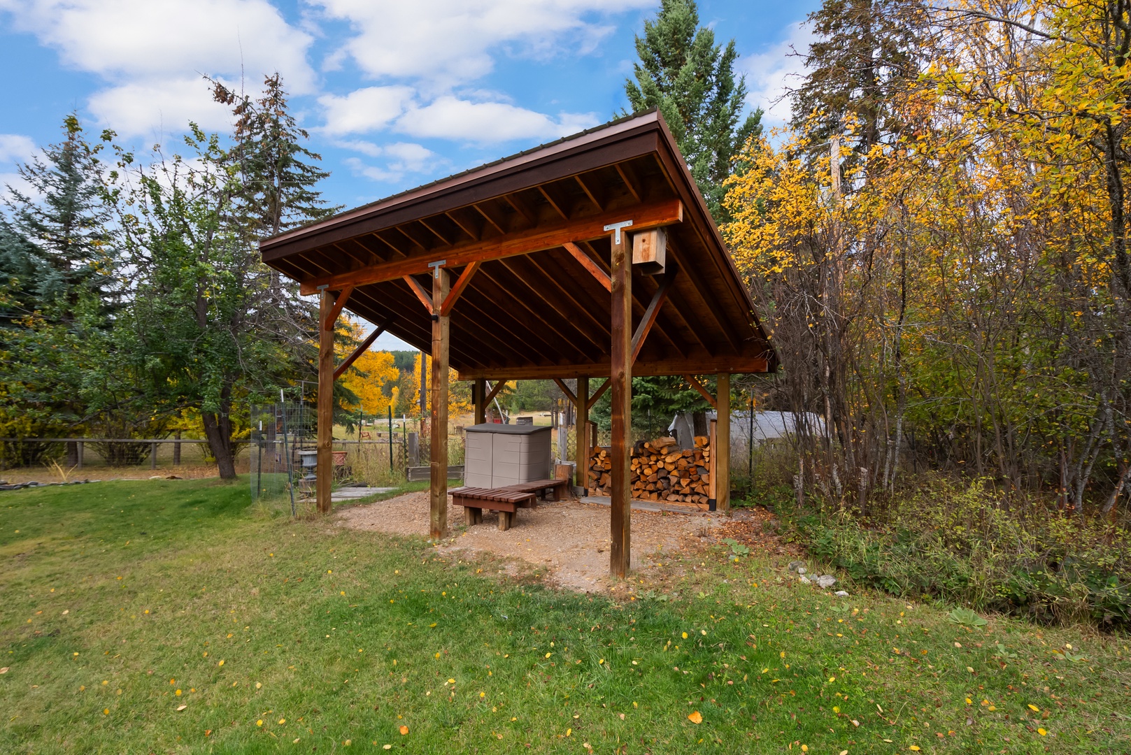 A covered outdoor pavilion nestled among autumn trees, featuring a hot tub and stacked firewood for cozy seasonal gatherings.