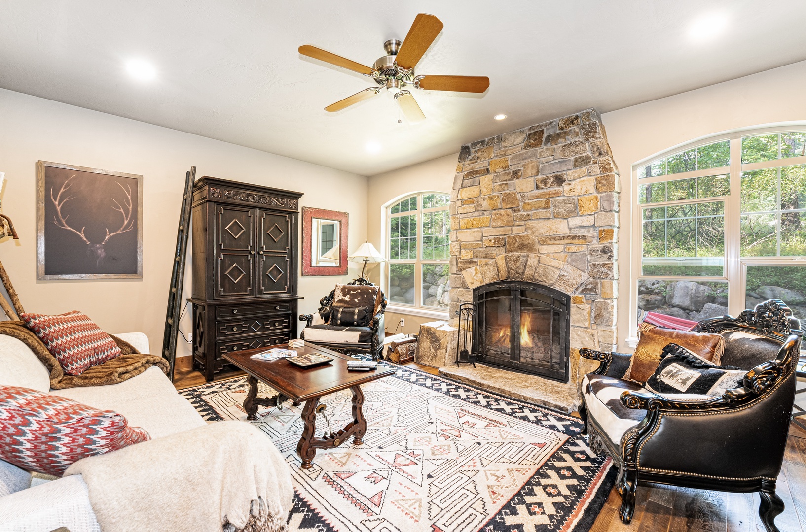 Gather with friends in your cozy living room, where a roaring stone fireplace and warm wooden accents invite relaxation. Large windows frame lush greenery outside, and rustic charm meets comfort in every corner.
