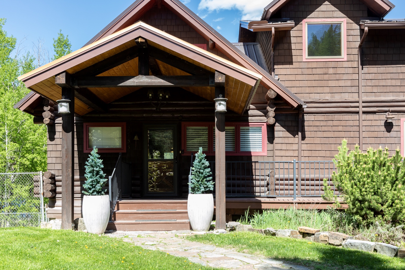 A charming mountain lodge-style entrance features rustic wood siding, a covered porch with wooden beams, and decorative planters with evergreen topiaries flanking the doorway. The natural stone walkway leads through a landscaped garden setting.