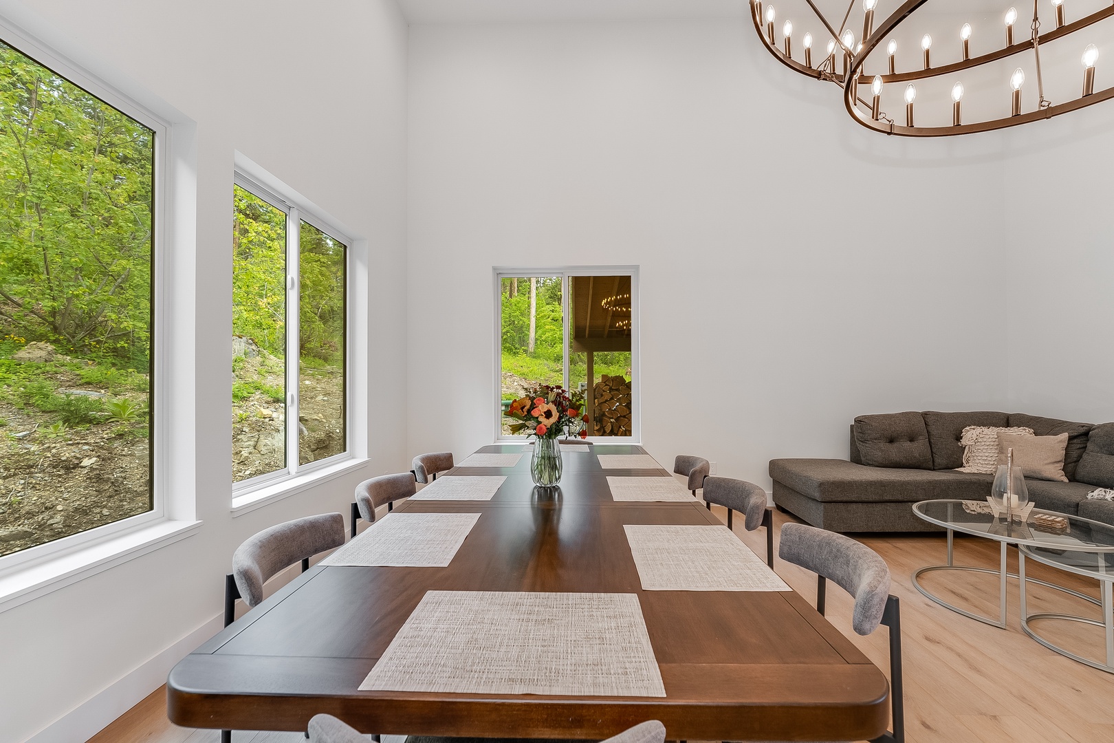 Gather around this elegant dining table where meals become memories. Natural light pours through floor-to-ceiling windows framing forest views, while the statement chandelier above creates a warm, inviting ambiance for your group.