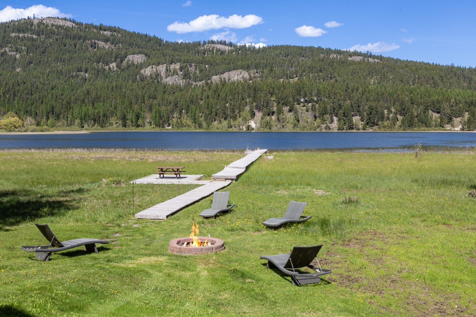 This lakefront property features a private dock, outdoor fire pit, and loungers, set against forested mountains and a tranquil blue lake, offering a peaceful natural retreat.