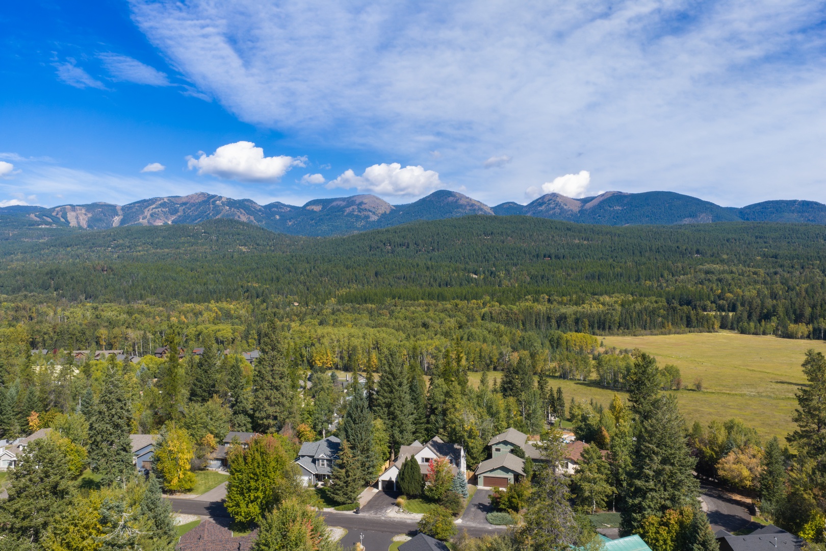 An aerial view captures a residential neighborhood nestled in a forested valley, with majestic mountains rising in the distance under expansive blue skies—a scenic mountain community setting.
