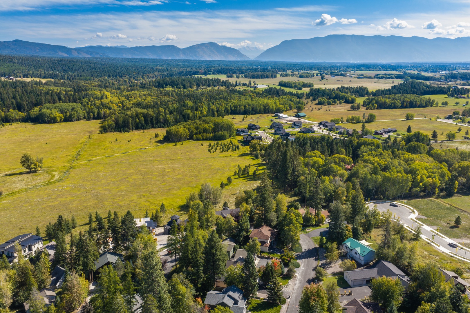 Aerial view captures the property nestled among evergreens, with sweeping mountain ranges and lush valley farmland stretching to the horizon beneath a bright summer sky.