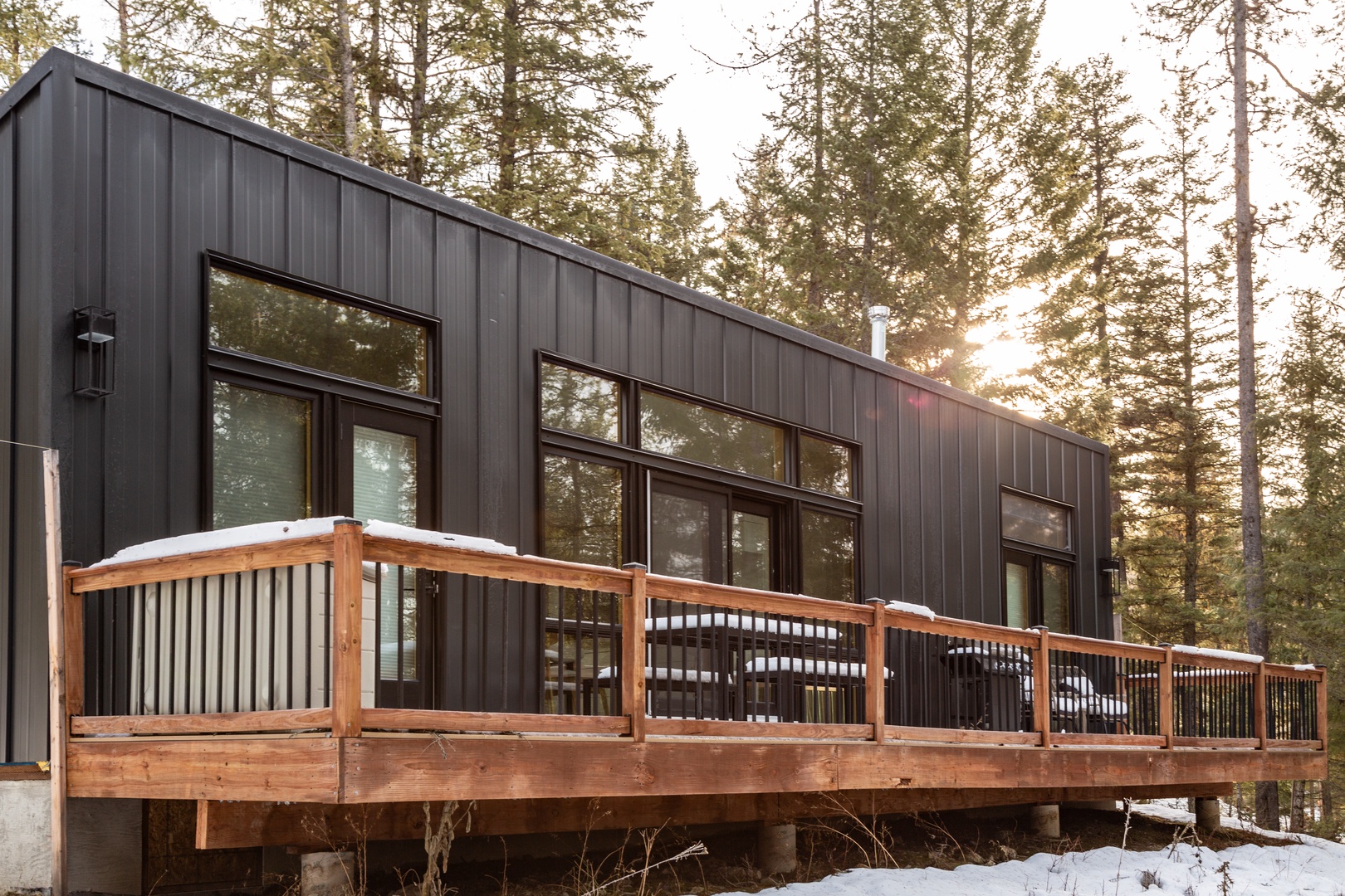 Modern vacation rental surrounded by tall evergreens and natural forest light, featuring a spacious wooden deck perfect for enjoying the peaceful winter setting.
