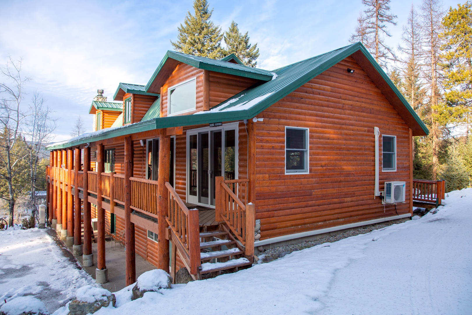 Charming log cabin with green metal roof and wraparound deck, nestled among evergreens in a peaceful winter setting—your mountain retreat awaits.