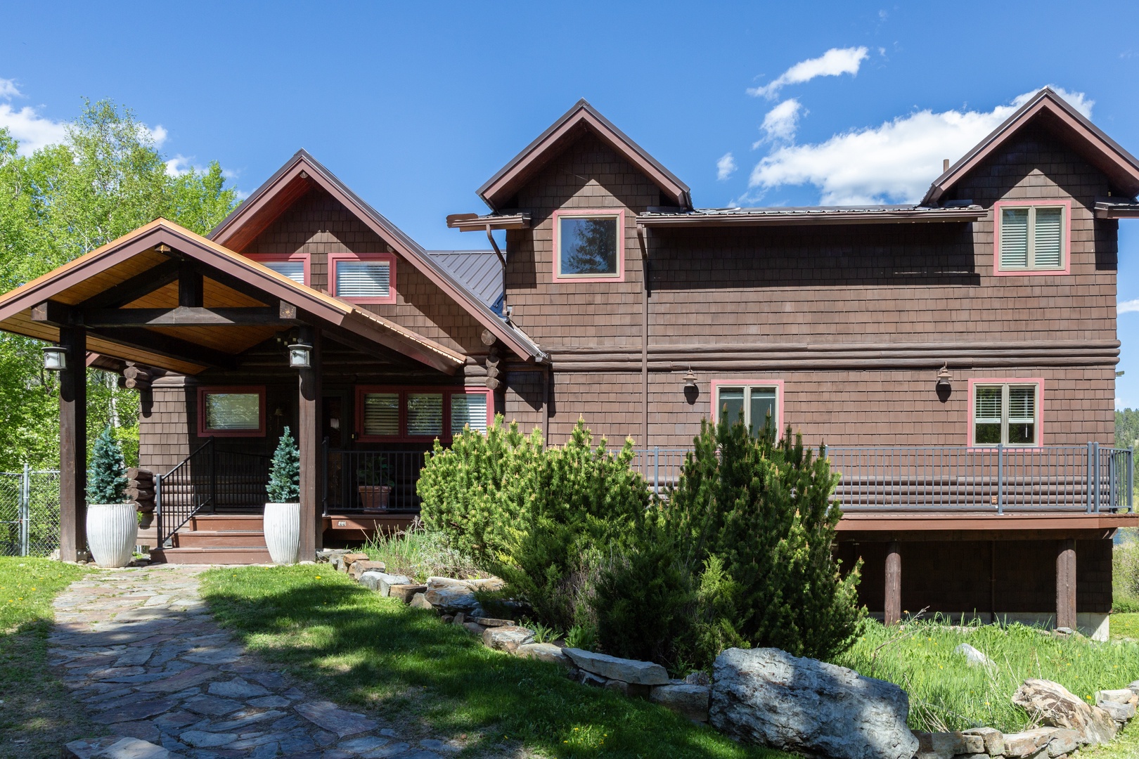 This charming mountain lodge features rustic elegance with distinctive pink-trimmed shingles and a welcoming covered entrance. Surrounded by lush greenery and natural landscaping, the property offers a peaceful retreat setting.