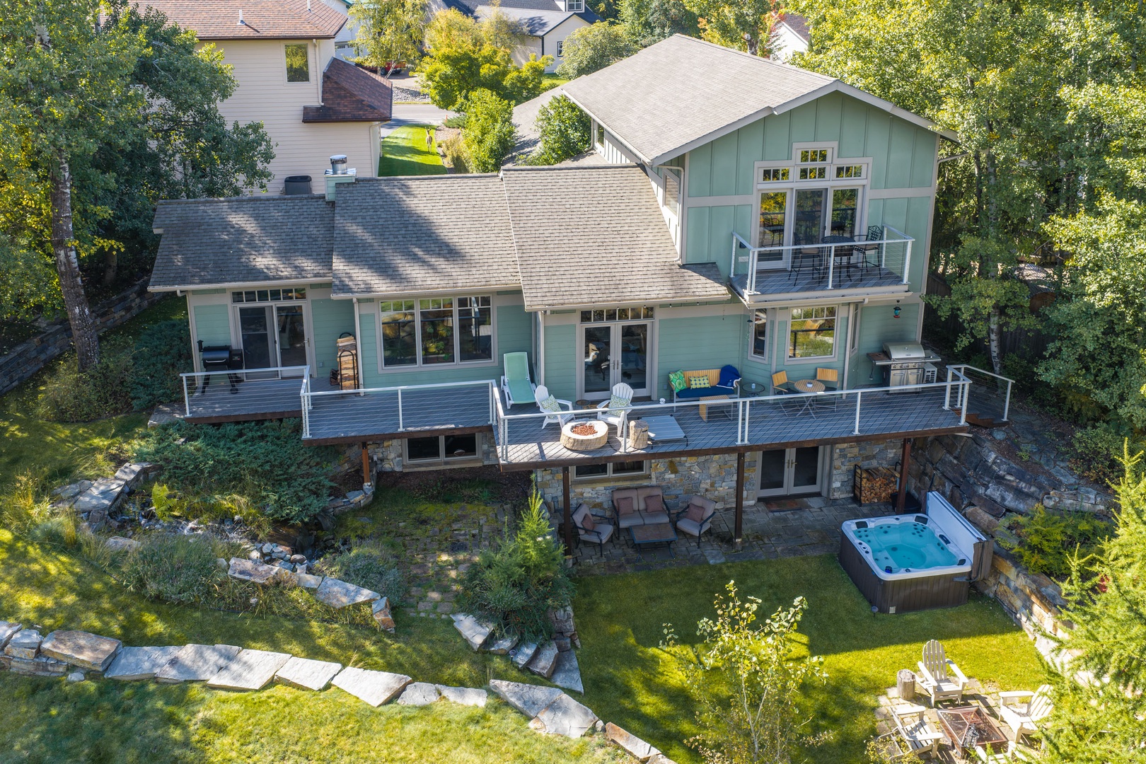 An aerial view showcasing the charming two-story rental property nestled in a peaceful, tree-lined neighborhood. The expansive deck, hot tub, and scenic surroundings create an ideal retreat for relaxation.