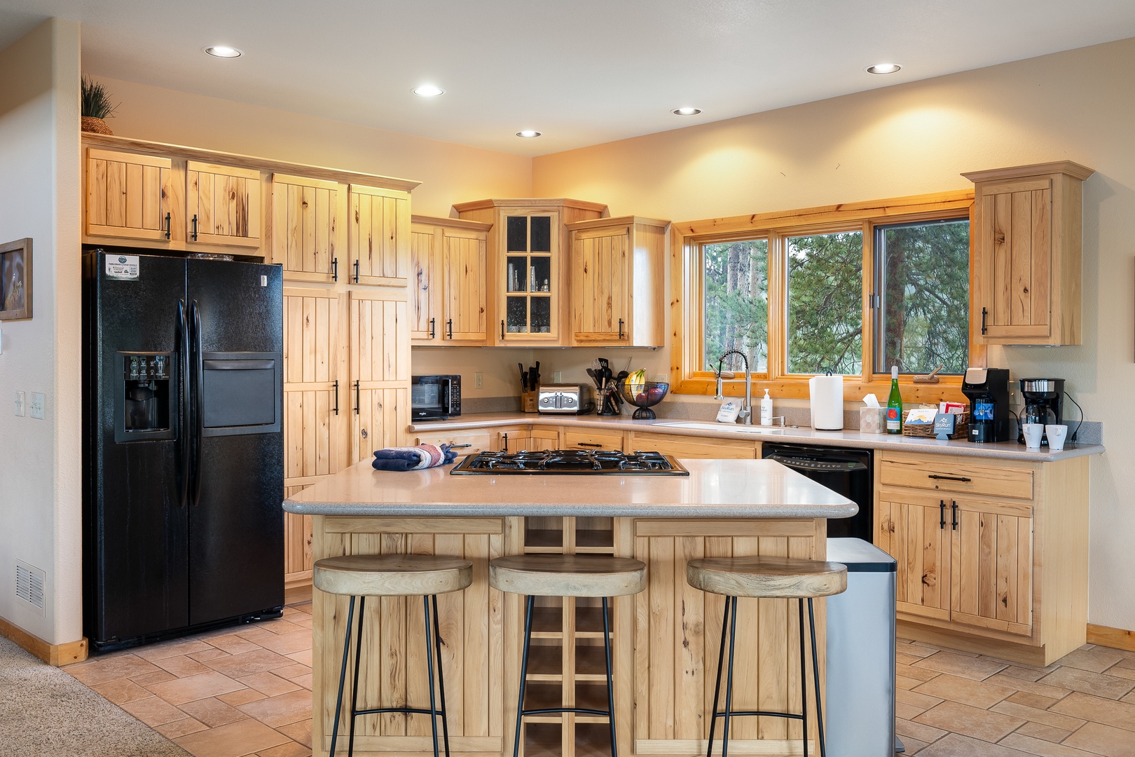 Your fully equipped kitchen features rustic wood cabinetry, modern appliances, and a spacious island with seating—perfect for preparing meals and gathering with loved ones as natural light streams through forest-facing windows.