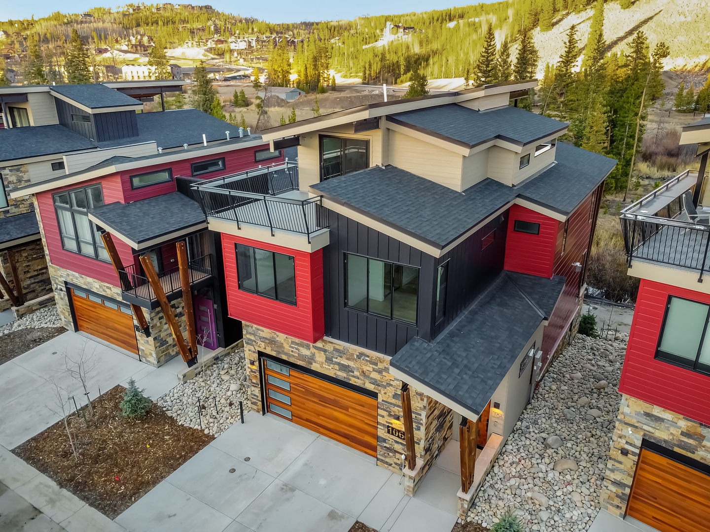 Modern mountain townhomes with striking red and stone facades nestled in a scenic alpine valley surrounded by forested hills.
