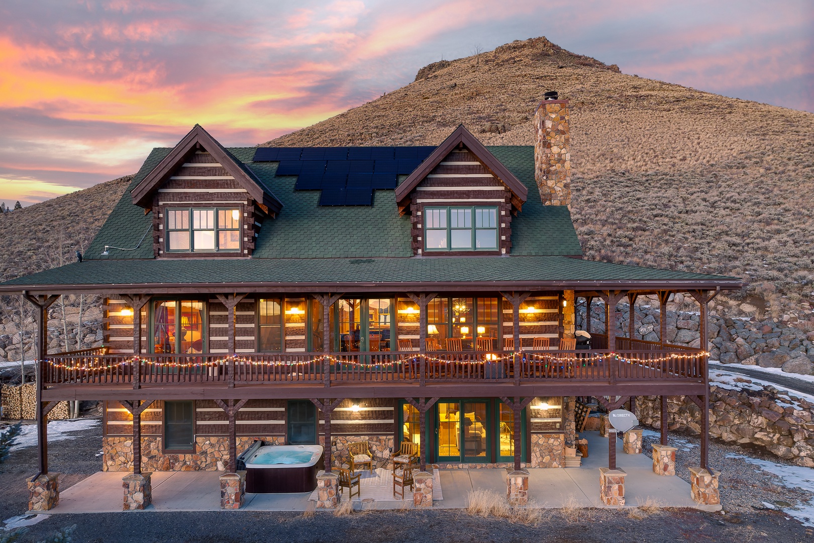 Rustic log cabin lodge nestled against rocky mountains, featuring stone and timber construction with wraparound decks and hot tub at sunset.