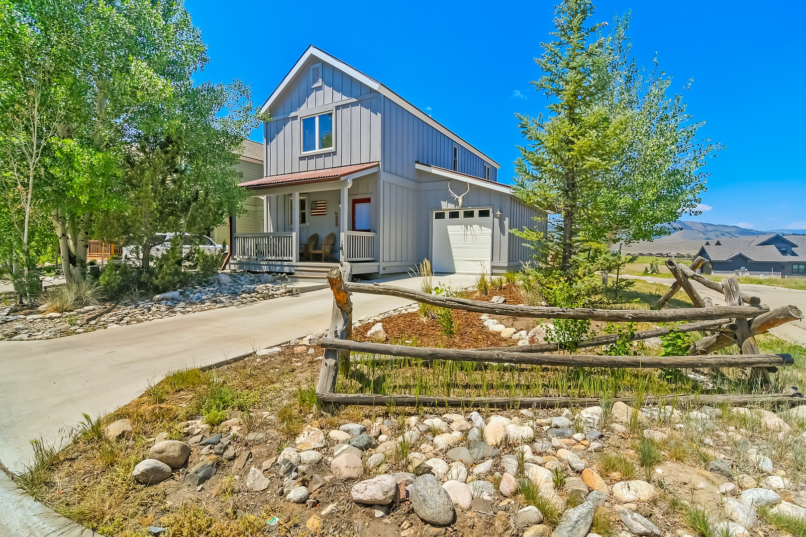Modern farmhouse-style vacation rental nestled in a serene mountain valley with rustic landscaping, covered front porch, and abundant natural light under bright blue skies.