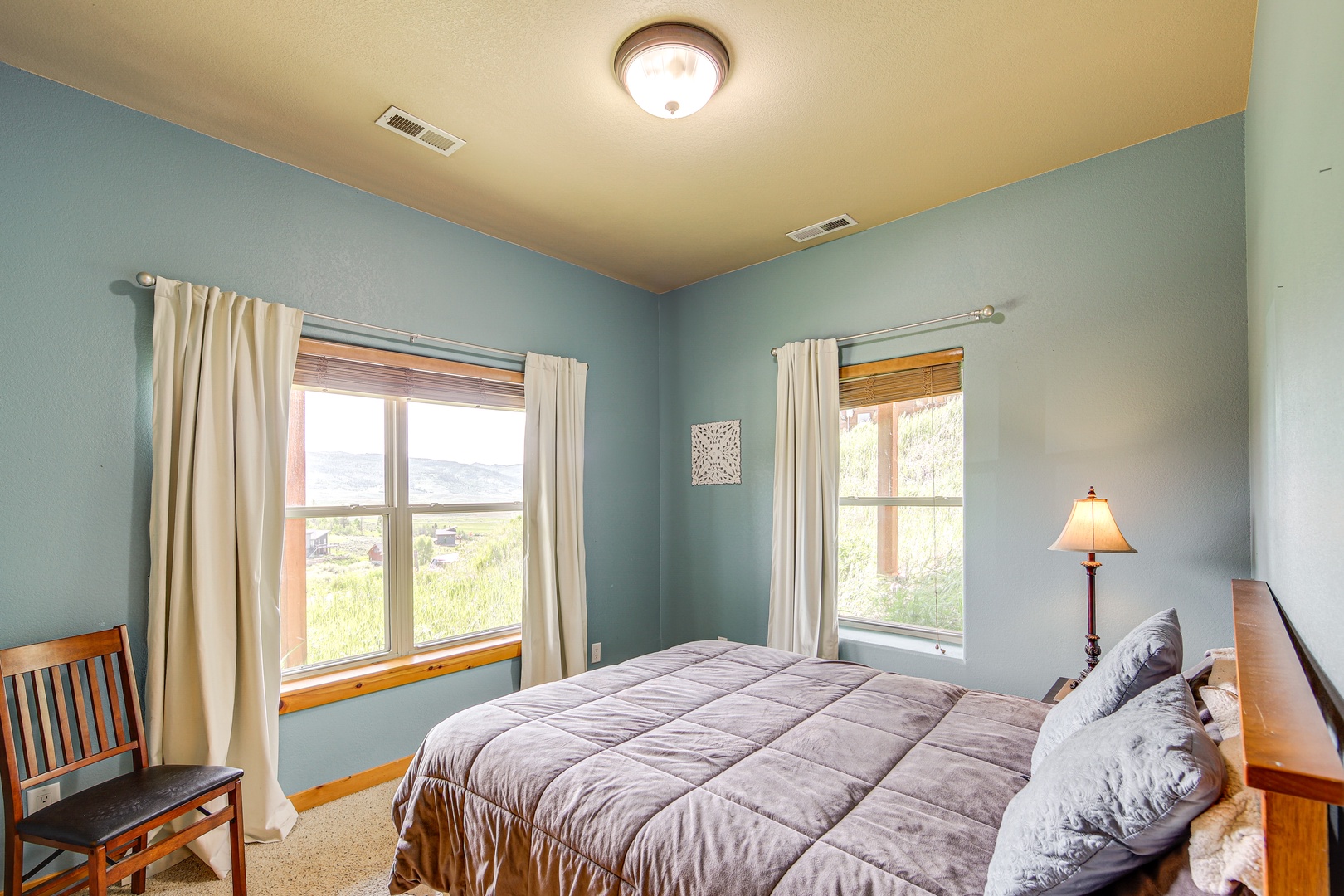 Your peaceful bedroom retreat features soothing blue walls and natural wood accents, with two large windows framing scenic views of rolling countryside.