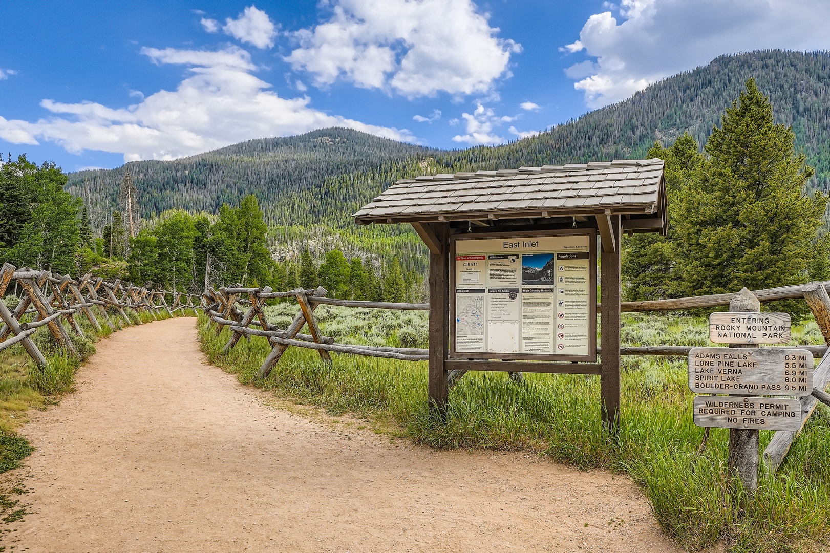 Trailhead entrance to Rocky Mountain National Park with hiking paths leading into forested mountains beneath a bright summer sky.