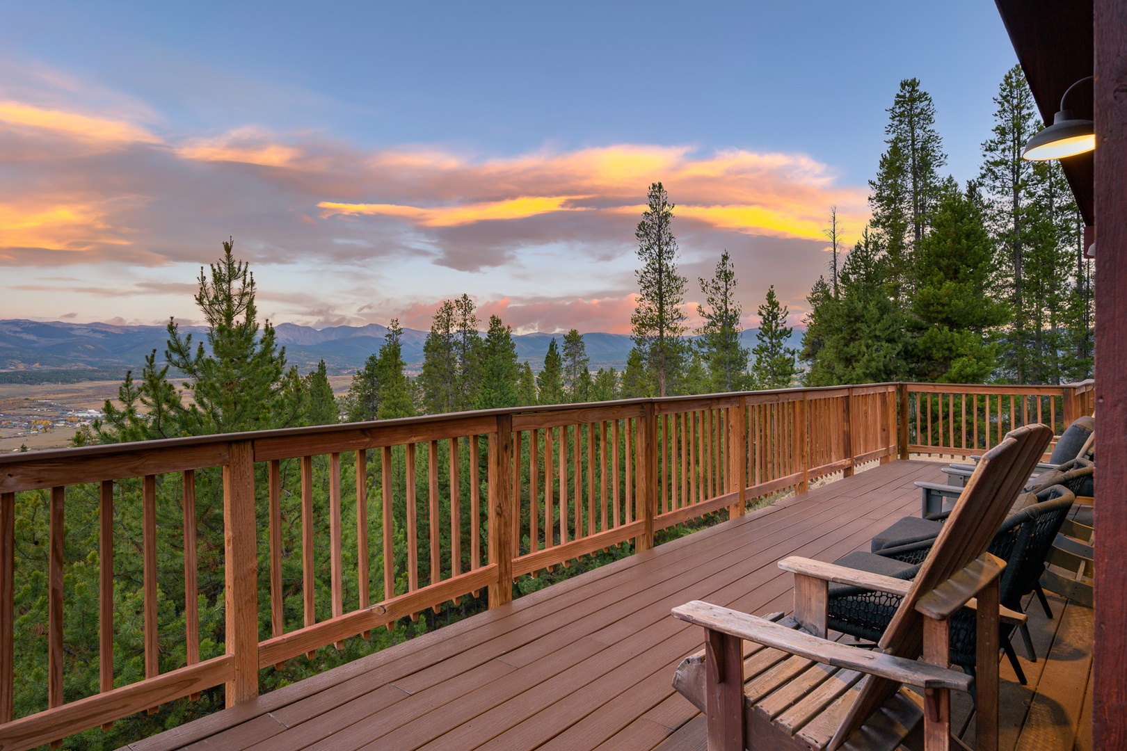 An expansive deck framed by towering pines opens onto distant mountain ridges bathed in golden sunset hues. This scenic terrace and surrounding natural landscape define the property's elevated hillside setting.