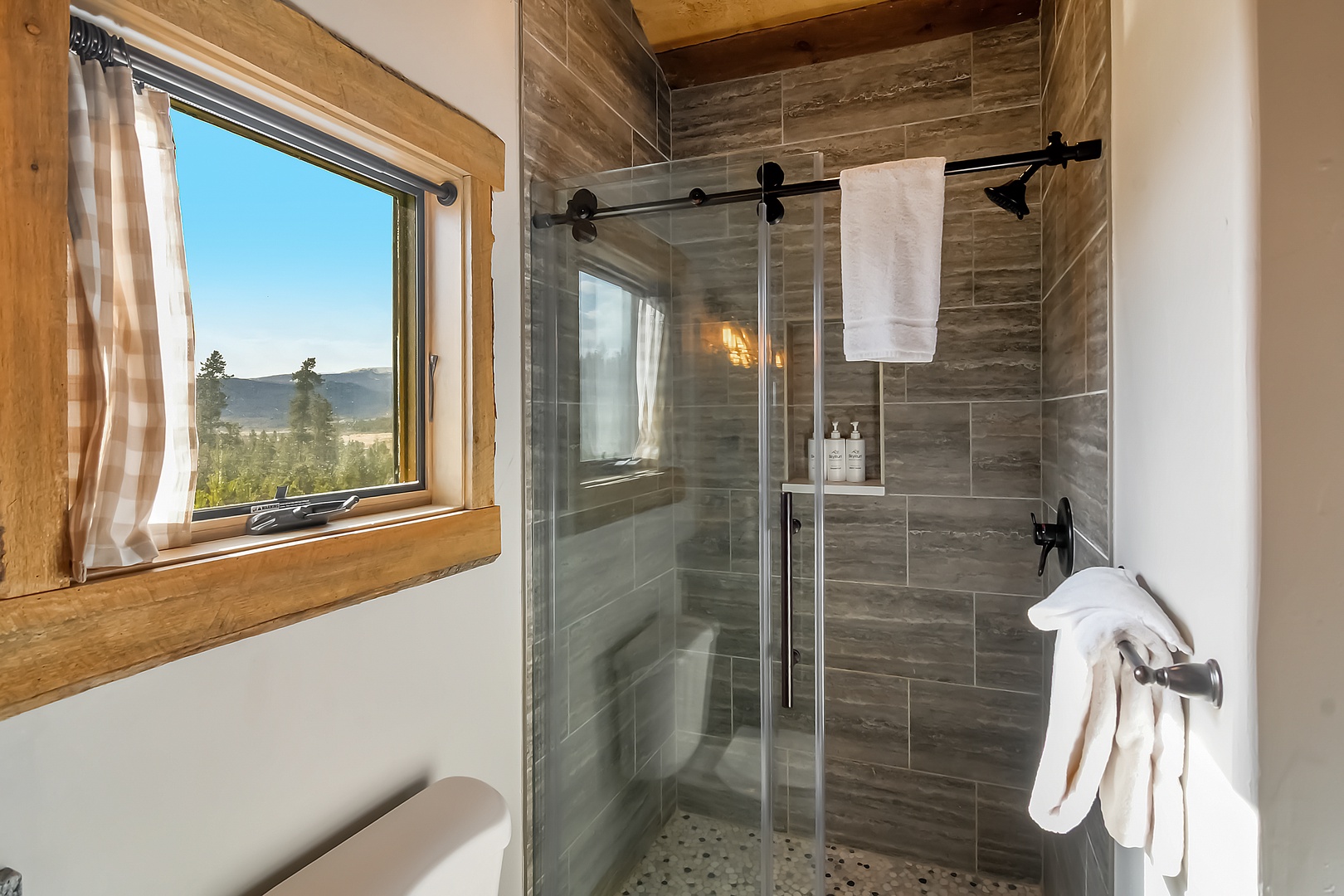 Refresh in your elegant shower with mountain views streaming through rustic wood-framed windows, surrounded by sophisticated gray stone tiles.