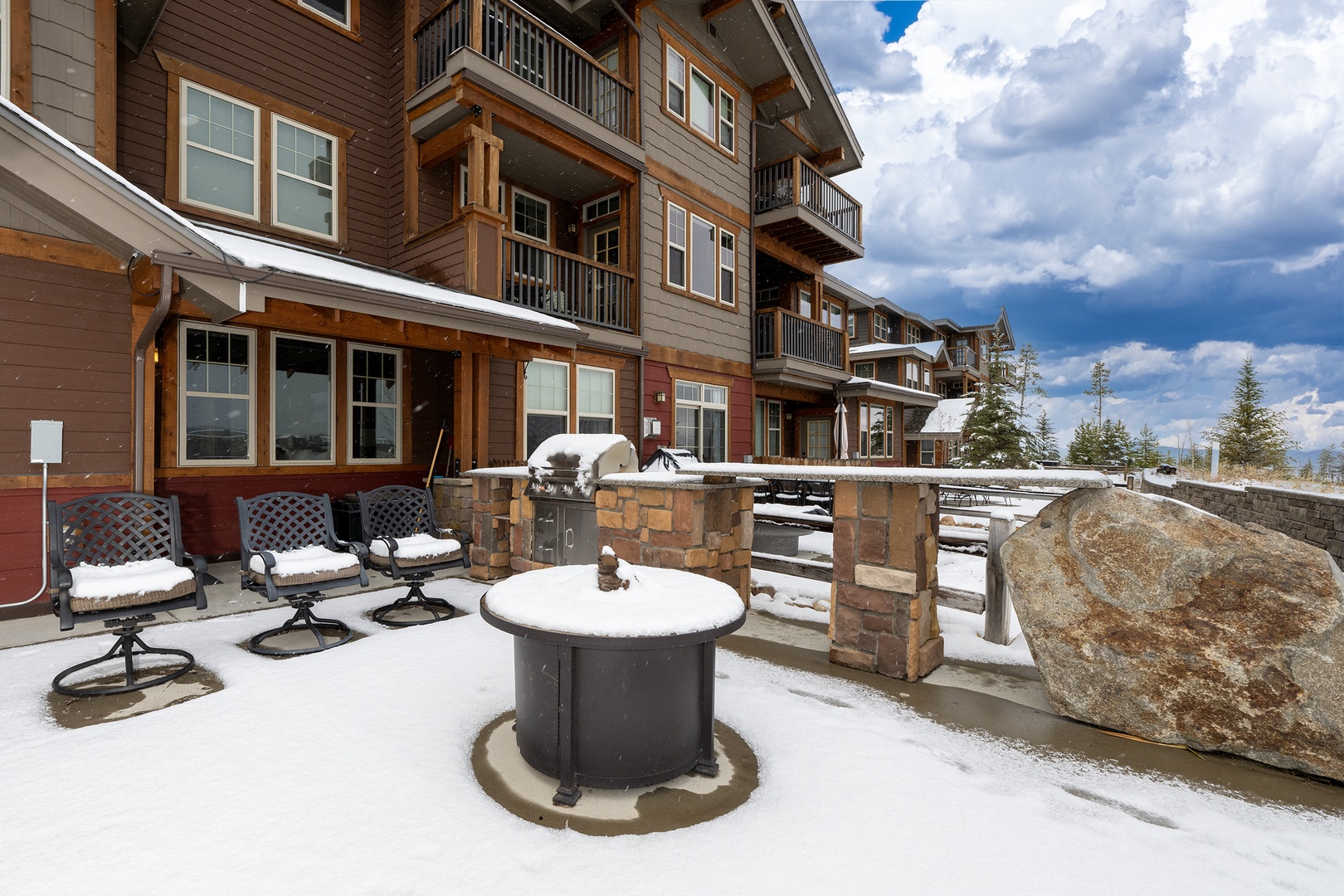 This multi-story mountain building features private patios with outdoor seating and BBQ facilities, nestled in a picturesque winter setting with dramatic cloudy skies and distant evergreen trees.
