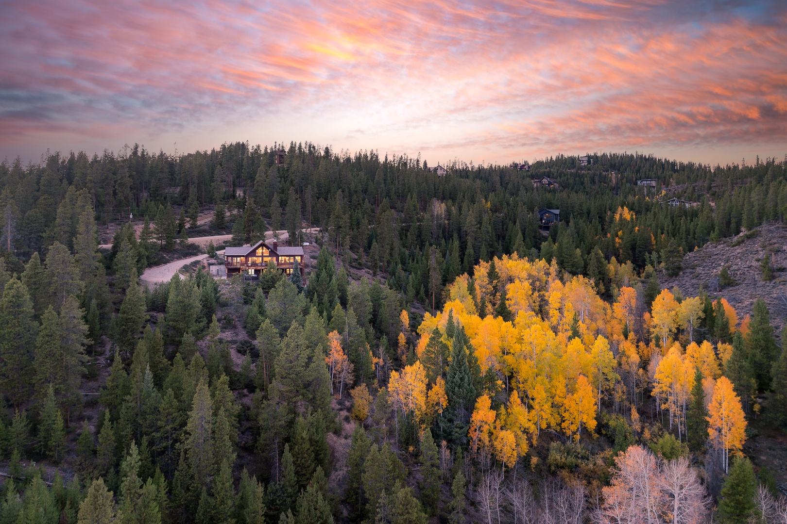 Aerial view of a mountain property nestled among evergreens and autumn aspens, captured at sunrise with dramatic skies painting the forest in golden light.