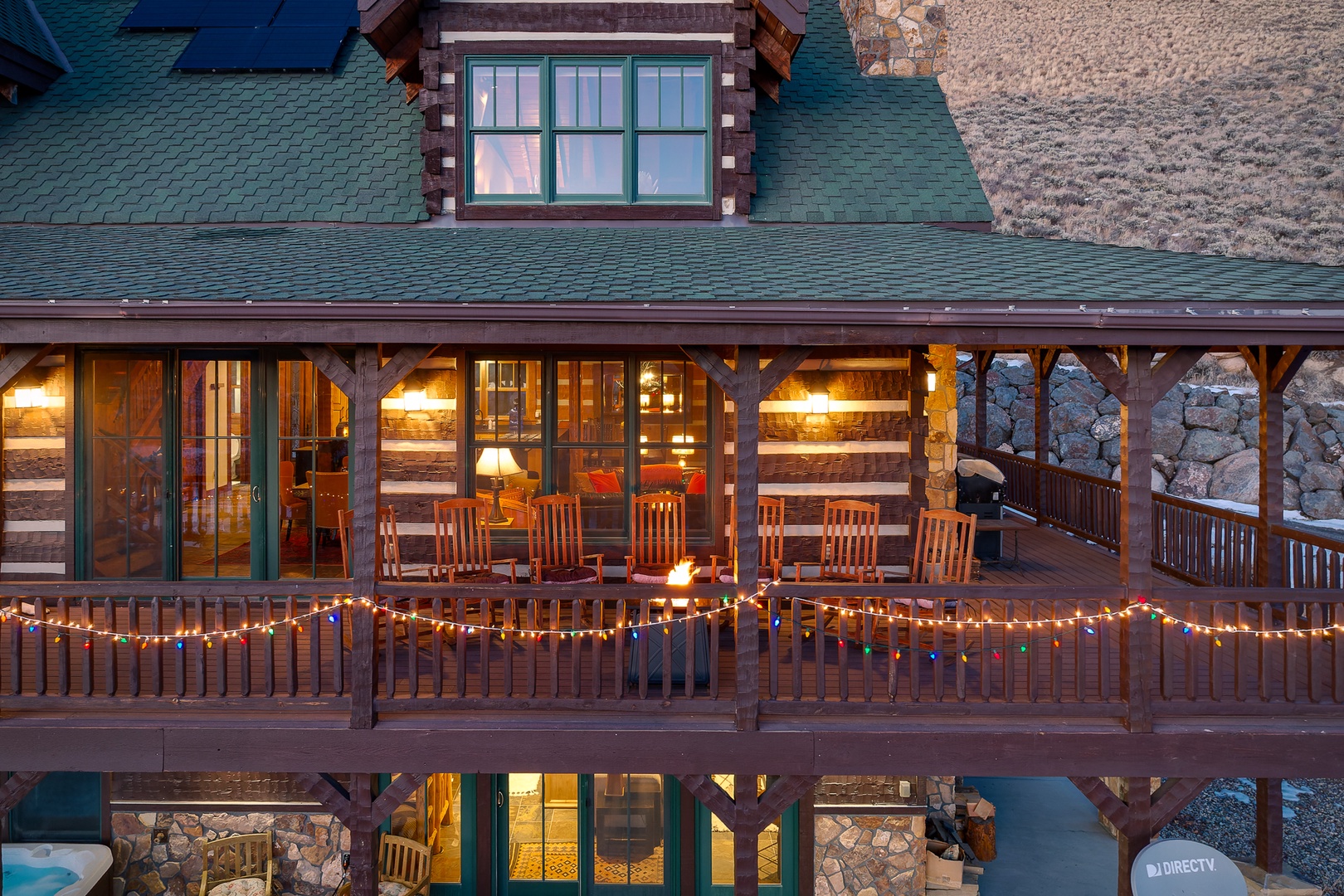 Rustic mountain cabin featuring a wrap-around deck with string lights, stone accents, and panoramic views of the surrounding natural landscape.