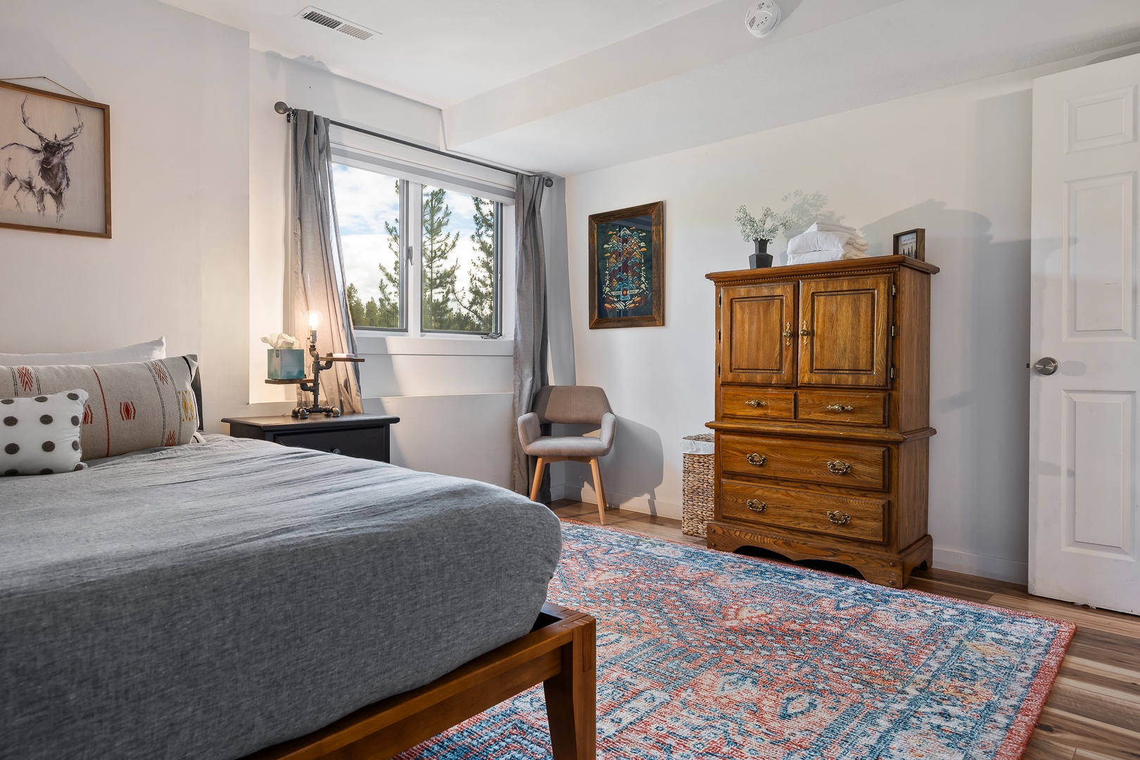Sunlight streams into your cozy bedroom, highlighting a plush bed, warm wood furnishings, and charming framed art. Wake refreshed with treetop views through soft gray curtains.