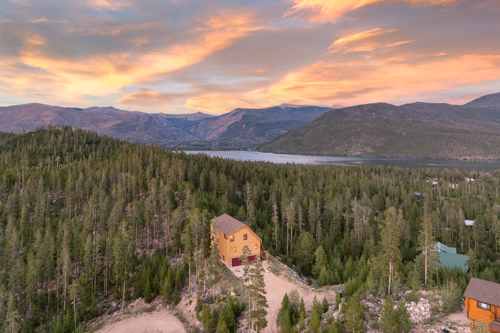 Nestled among evergreens with mountain peaks and a shimmering lake beyond, this property sits in a stunning natural setting where vibrant sunset skies paint the horizon.