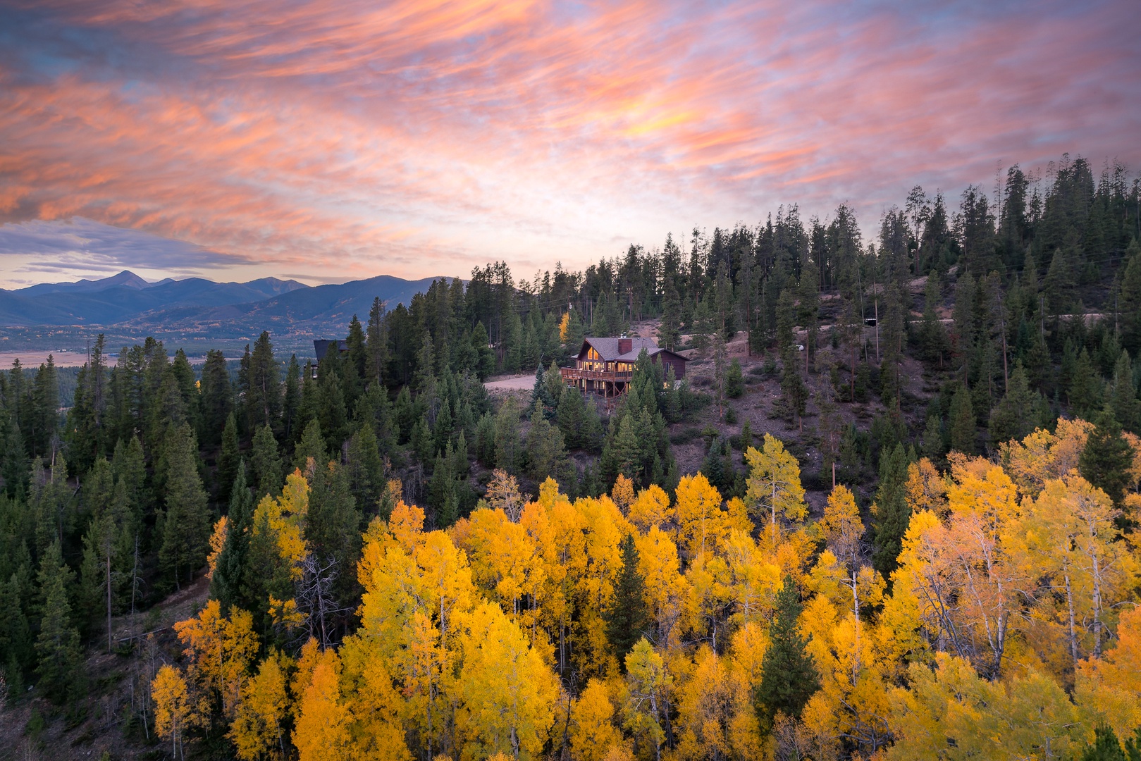 A vacation rental nestled in forested hills, surrounded by golden aspens and evergreens, with mountains visible in the distance under a vibrant sunset sky.